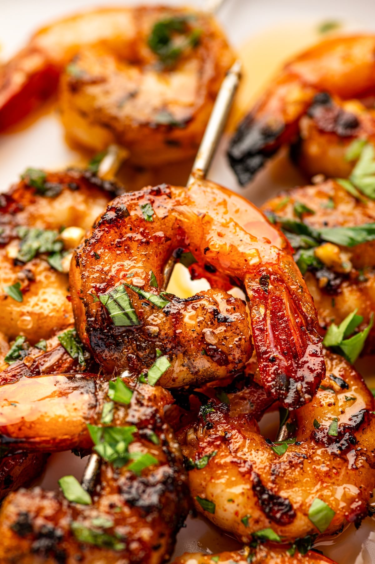 Extreme close-up of juicy grilled shrimp with dark char marks, chopped garlic, black pepper, and fresh parsley. The shrimp are threaded onto metal skewers and coated in a glossy lemon garlic marinade.