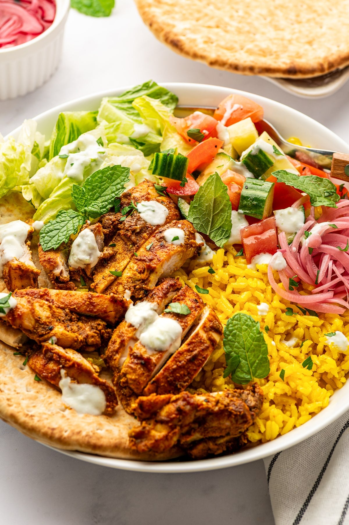 A large white bowl filled with sliced chicken shawarma, bright yellow rice, chopped romaine, cucumber tomato salad, pink pickled onions, creamy white tzatziki, mint, parsley, and pita, with extra pita visible above the bowl.