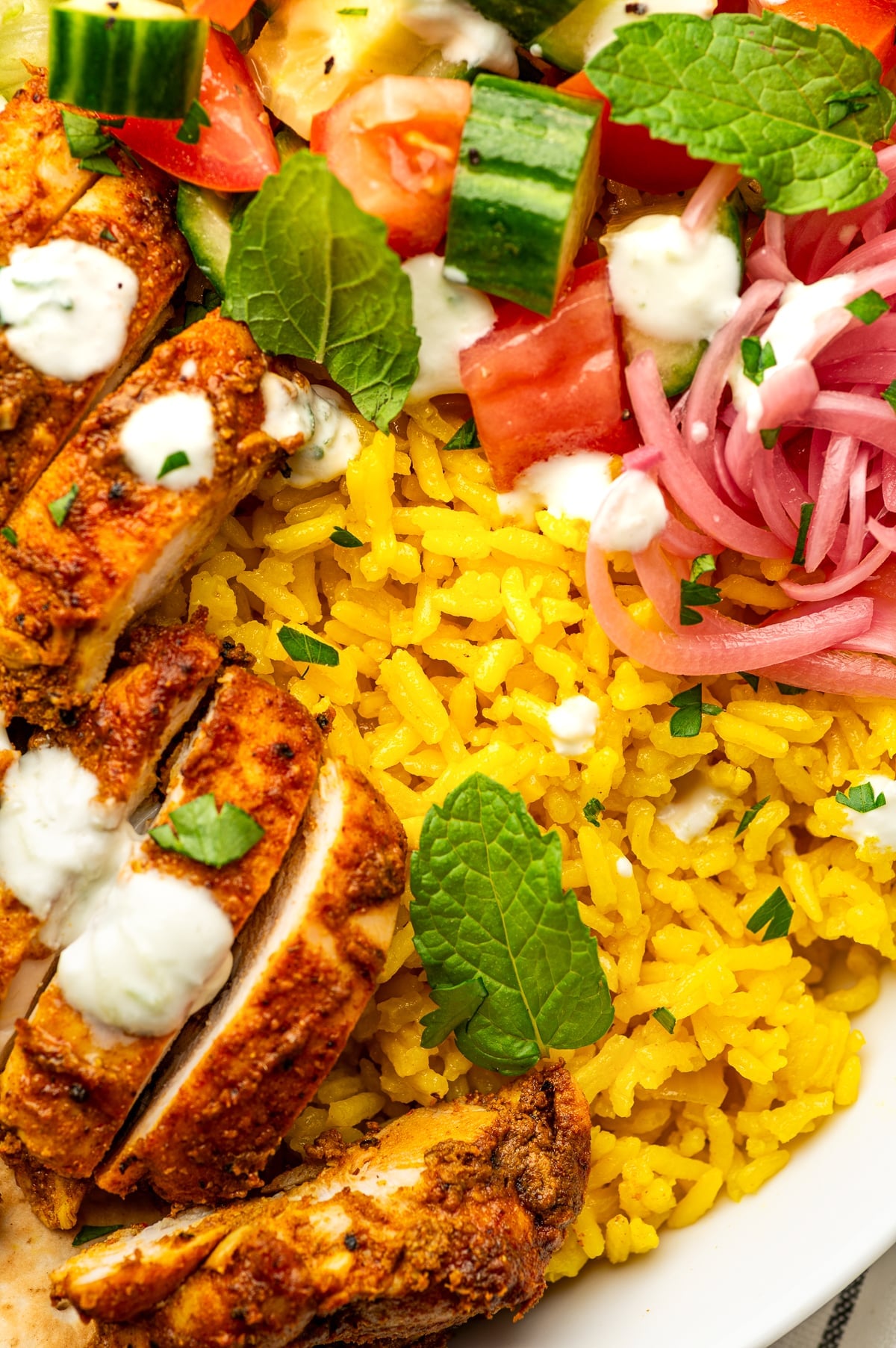 Close-up of bright golden yellow rice in a white chicken shawarma bowl with sliced browned chicken, creamy white tzatziki, pink pickled red onions, chopped cucumber and tomato, parsley, and fresh green mint leaves.