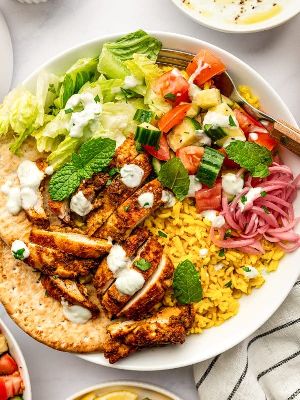 A large white chicken shawarma bowl filled with sliced spiced chicken, golden turmeric rice, chopped romaine, cucumber tomato salad, pink pickled onions, creamy tzatziki, fresh mint, parsley, and pita, surrounded by lemon wedges and small side dishes.