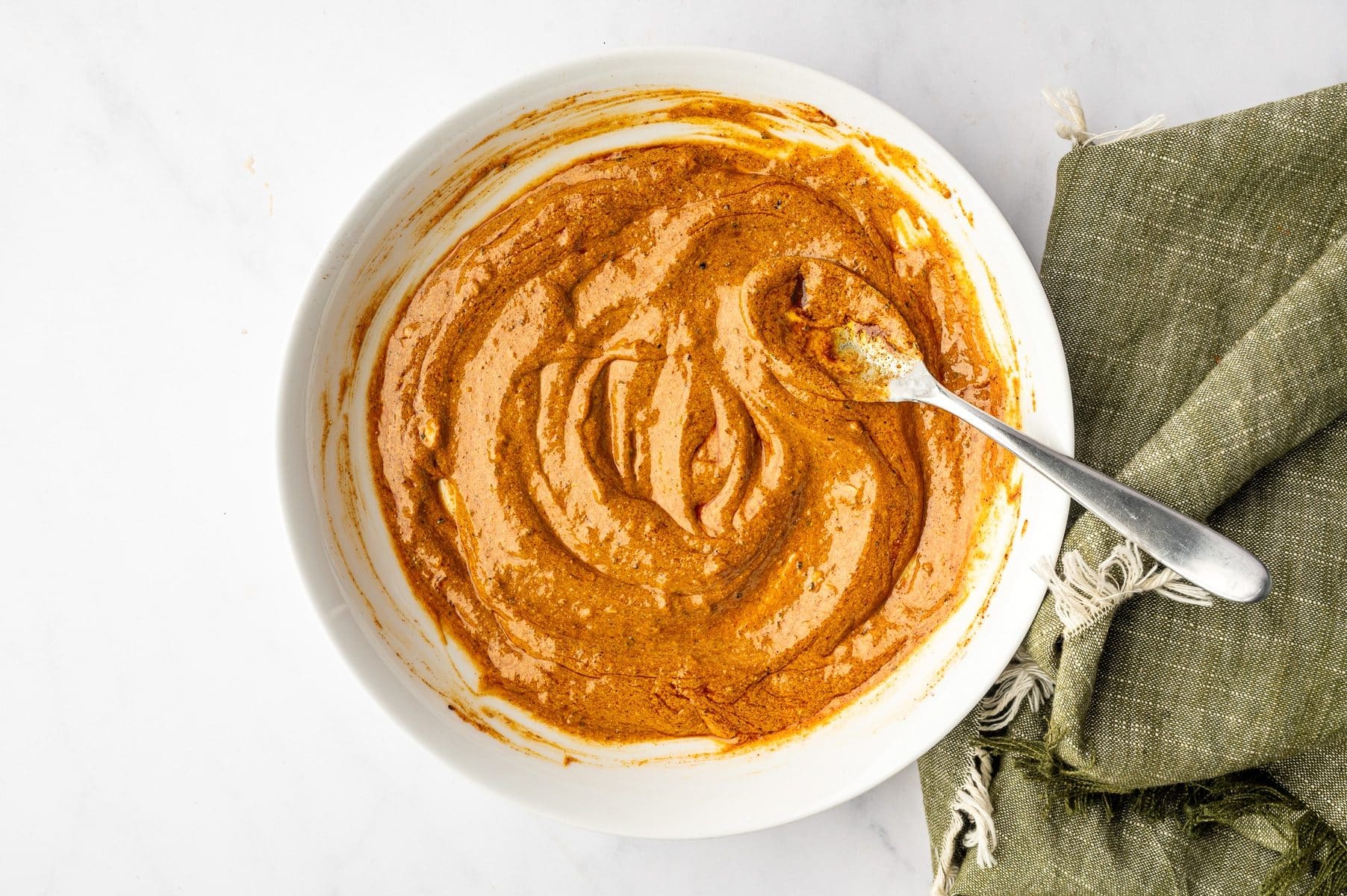 Thick orange shawarma marinade swirled inside a large white ceramic bowl with a silver spoon, set beside an olive-green fringed napkin on a white marble surface.