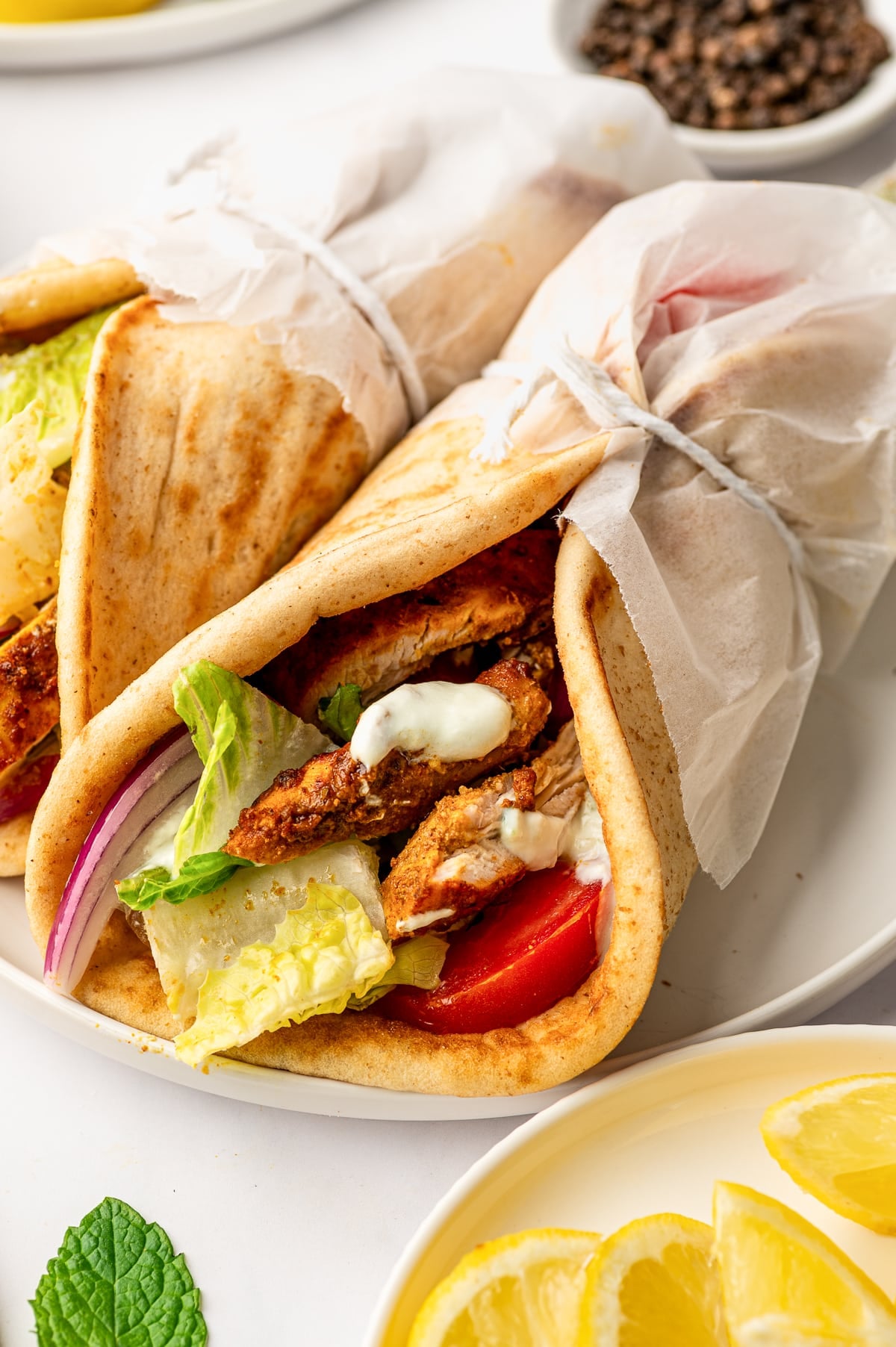 Close-up of a tan pita wrap filled with sliced orange-red chicken shawarma, green romaine lettuce, red tomato, purple-red onion, and white tzatziki sauce, with two parchment-wrapped pitas behind it and yellow lemon slices in the foreground.