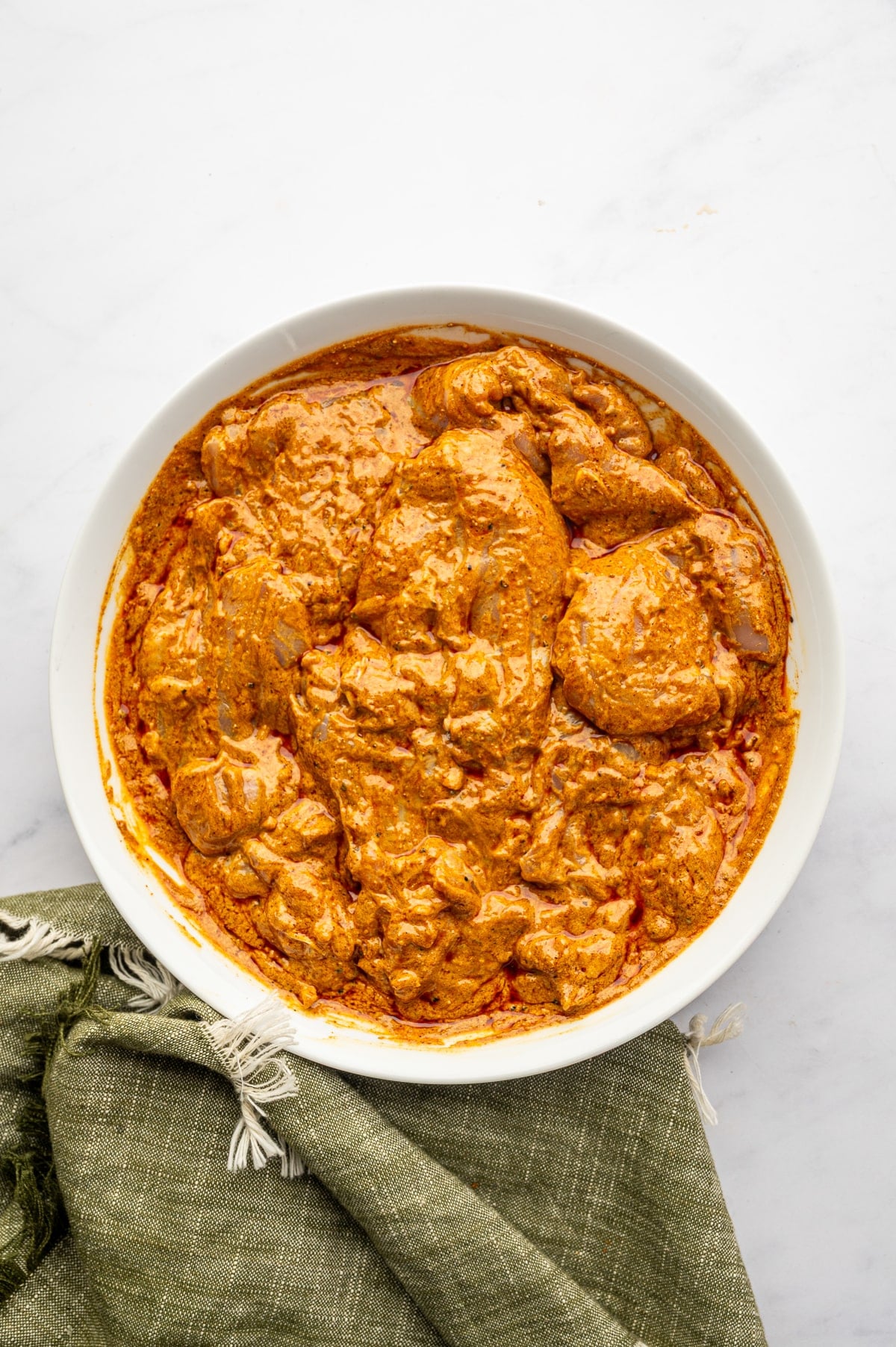 Raw boneless, skinless chicken thighs thoroughly coated in orange yogurt shawarma marinade inside a large white ceramic bowl, with an olive-green fringed napkin at the lower left.