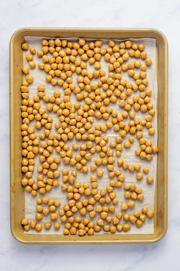 Canned chickpeas lightly coated with olive oil and salt and spread in a single layer on white parchment paper inside a gold-colored rimmed baking sheet before roasting.
