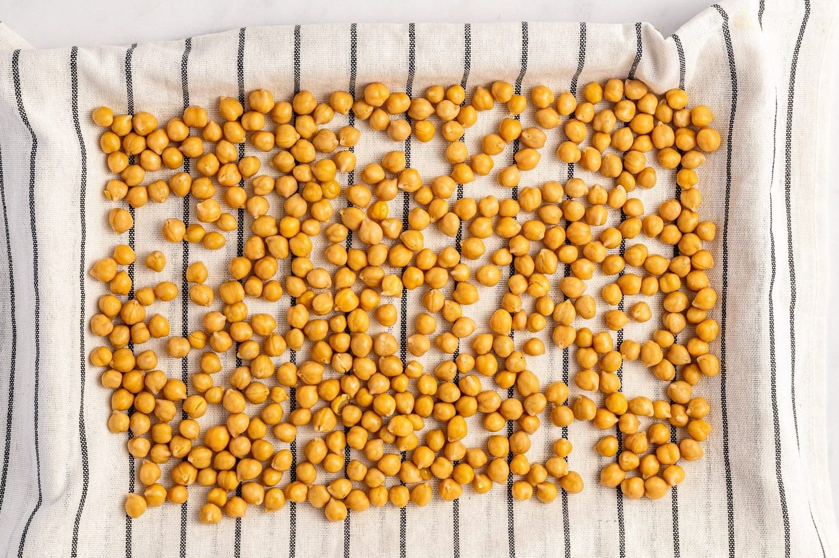 Drained canned chickpeas spread in a single layer over a white kitchen towel with thin black stripes to dry before roasting, photographed overhead on a white surface.