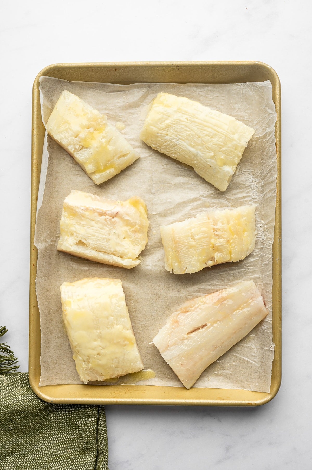 Six raw cod fillets of varying sizes arranged on tan parchment paper inside a gold rimmed baking sheet and brushed with melted butter. An olive-green cloth napkin is partially visible in the lower-left corner.