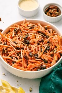 Close-up of creamy carrot raisin salad in a white serving bowl with shredded carrots, raisins, chopped pecans, black pepper, lemon wedges, and a green napkin nearby.