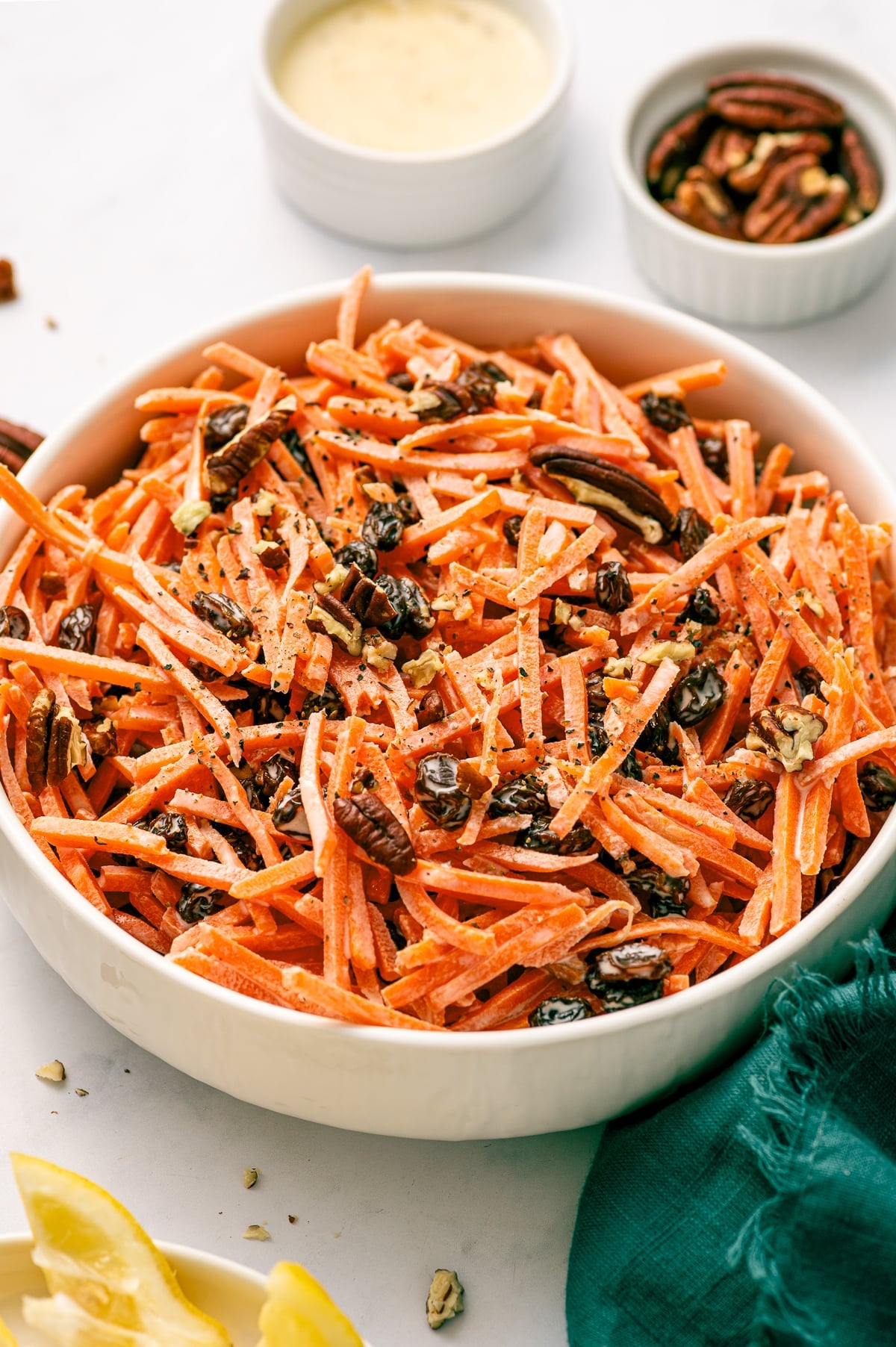 Close-up of creamy carrot raisin salad in a white serving bowl with shredded carrots, raisins, chopped pecans, black pepper, lemon wedges, and a green napkin nearby.