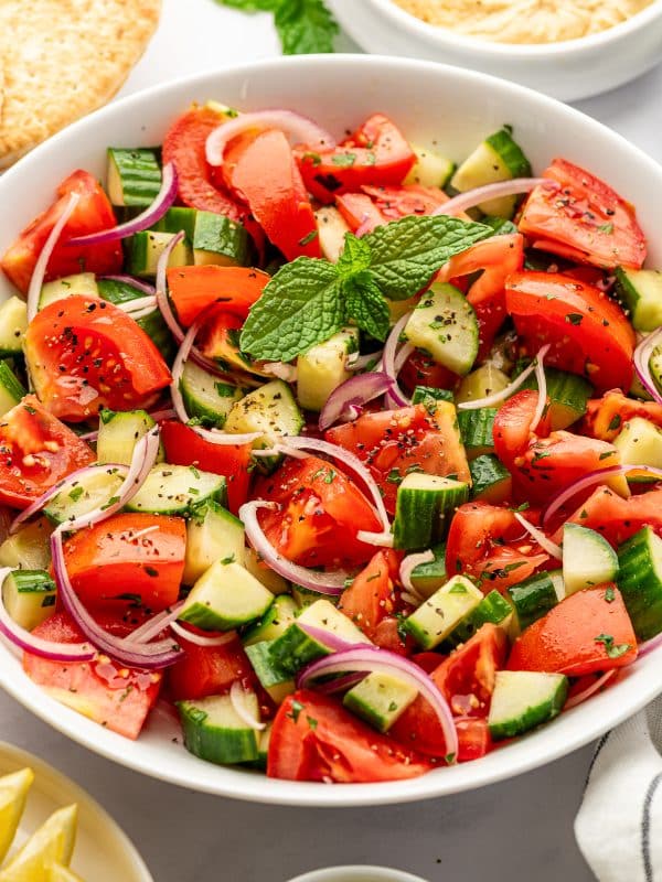 Close-up of cucumber tomato salad in a white serving bowl filled with juicy red tomato wedges, chunky green cucumber pieces, thinly sliced purple-red onion, chopped parsley, black pepper, and a bright green mint sprig, with pita bread and hummus in the background.