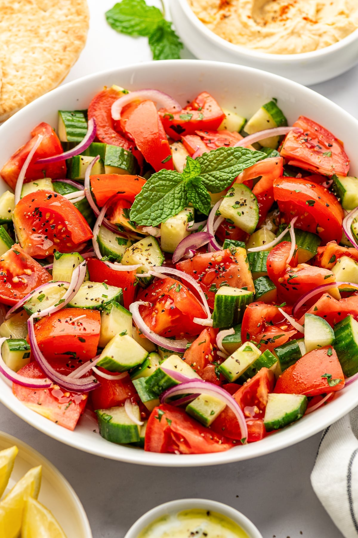 Close-up of cucumber tomato salad in a white serving bowl filled with juicy red tomato wedges, chunky green cucumber pieces, thinly sliced purple-red onion, chopped parsley, black pepper, and a bright green mint sprig, with pita bread and hummus in the background.