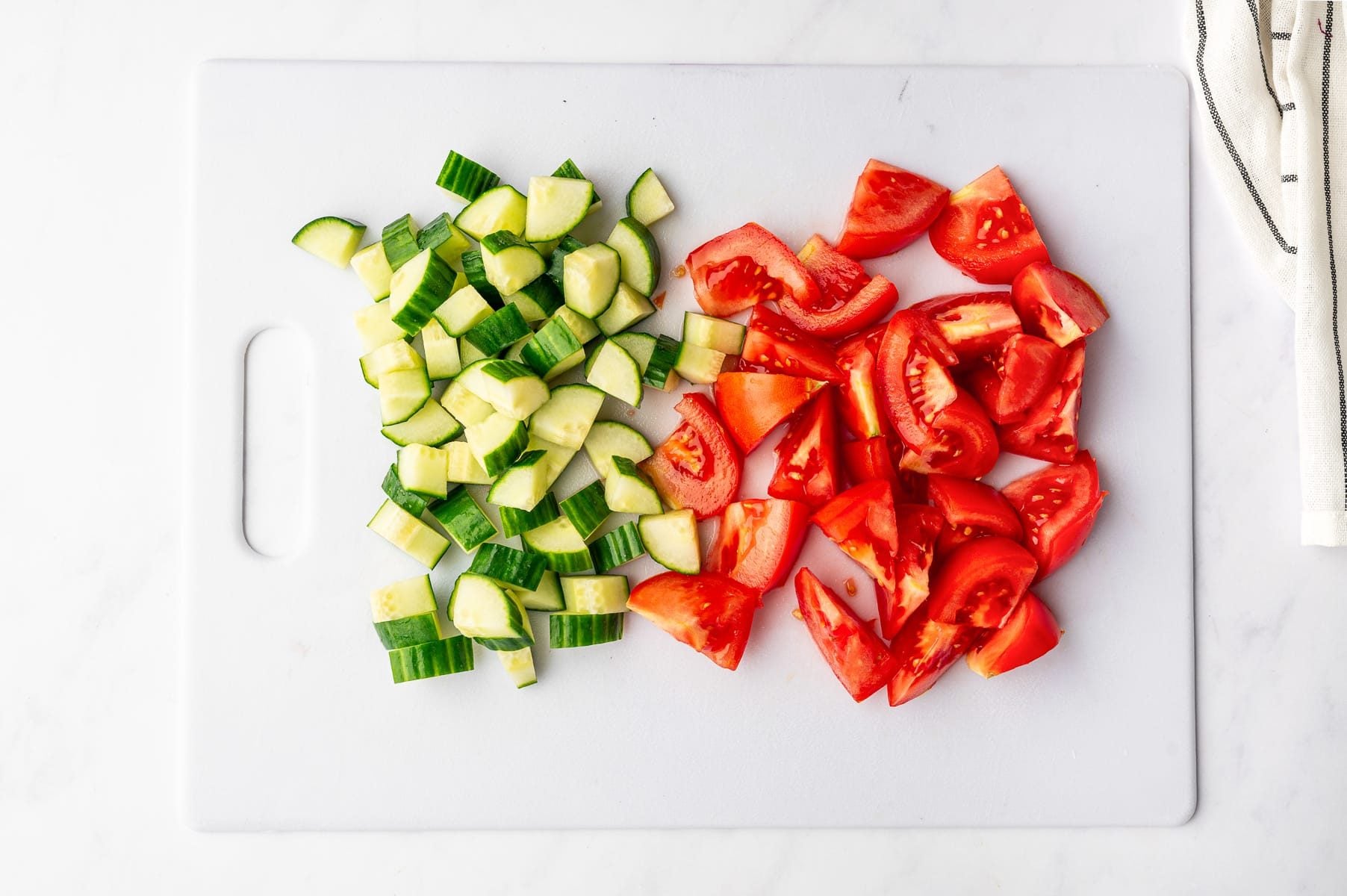 Chopped English cucumber and ripe red tomatoes arranged separately on a large white cutting board, with a folded white and black striped kitchen towel along the bottom edge.