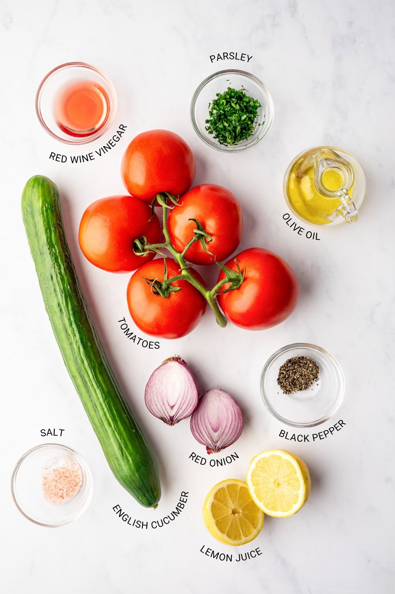 Labeled cucumber tomato salad ingredients arranged on a white marble surface, including red tomatoes on the vine, a green English cucumber, halved red onion, chopped parsley, lemon halves, olive oil, red wine vinegar, salt, and black pepper, with black text identifying each ingredient.