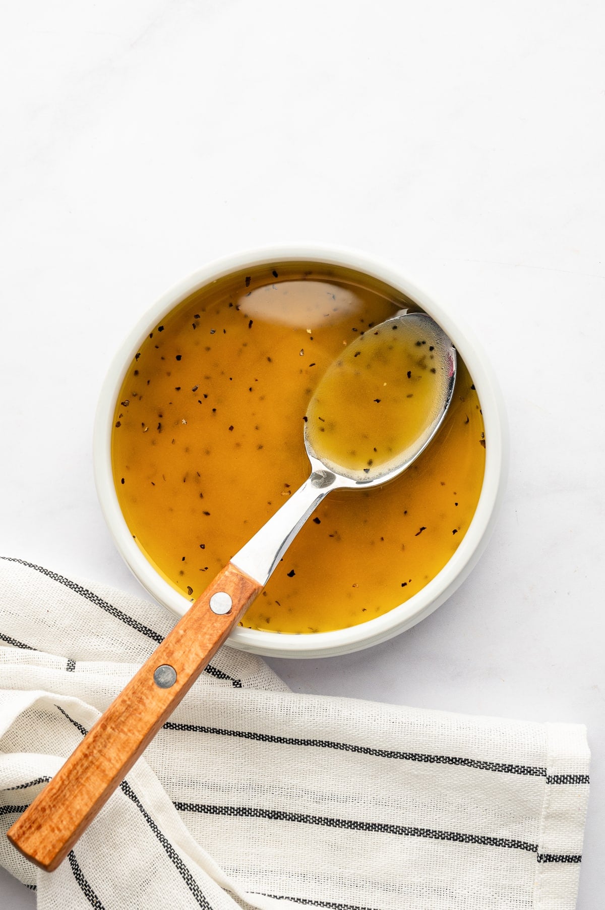 Golden lemon and red wine vinaigrette flecked with black pepper in a shallow white bowl, with a silver spoon featuring a wooden handle resting inside and a striped kitchen towel underneath.