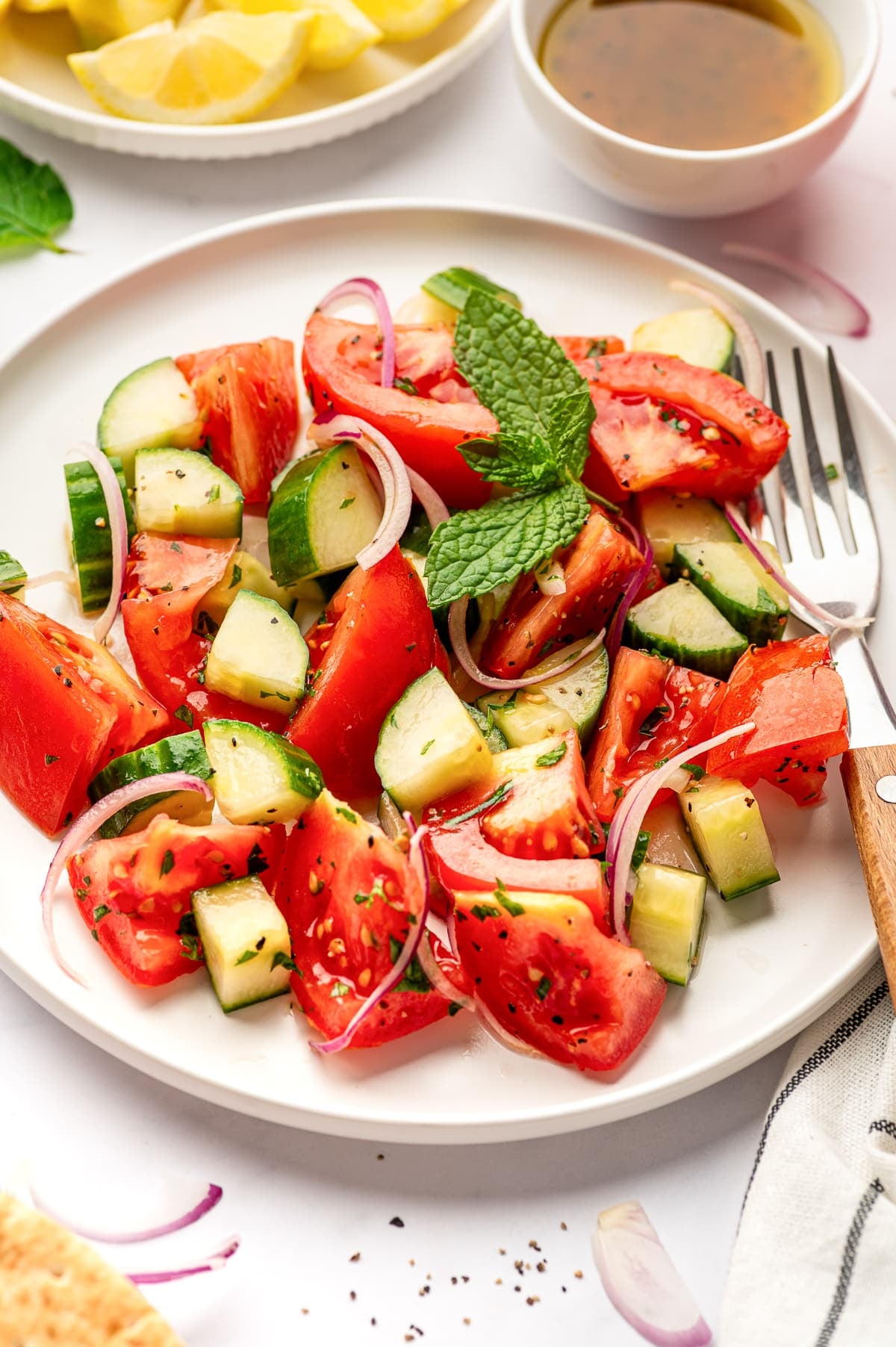 Cucumber tomato salad served on a round white plate with red tomato wedges, chopped green cucumber, thinly sliced red onion, parsley, black pepper, and a fresh mint garnish, alongside a silver fork with a wooden handle, lemon wedges, pita bread, and a small white bowl of vinaigrette.