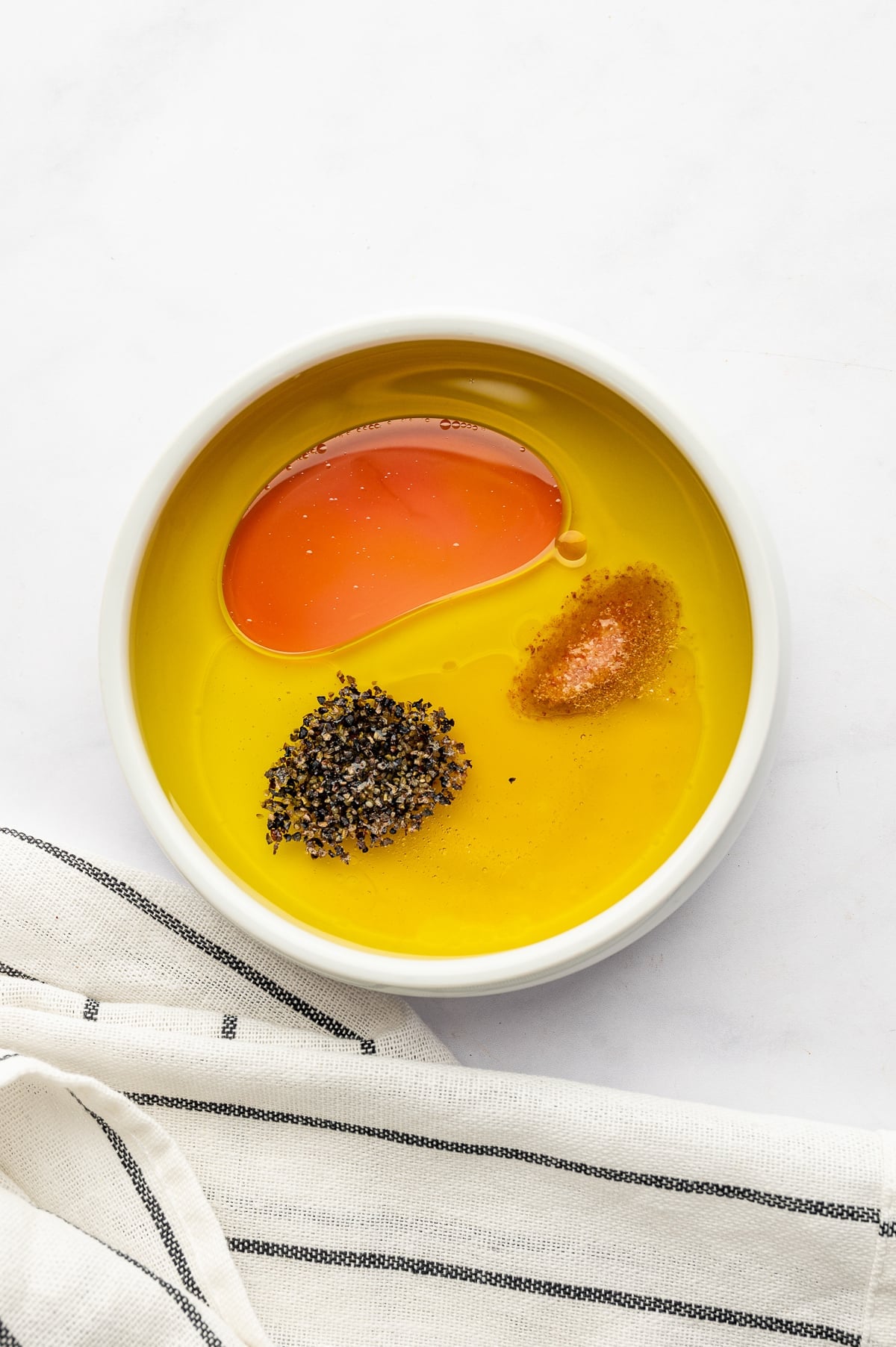 Olive oil, red wine vinegar, salt, and coarse black pepper arranged separately in a shallow white bowl, with a white and black striped kitchen towel draped underneath.