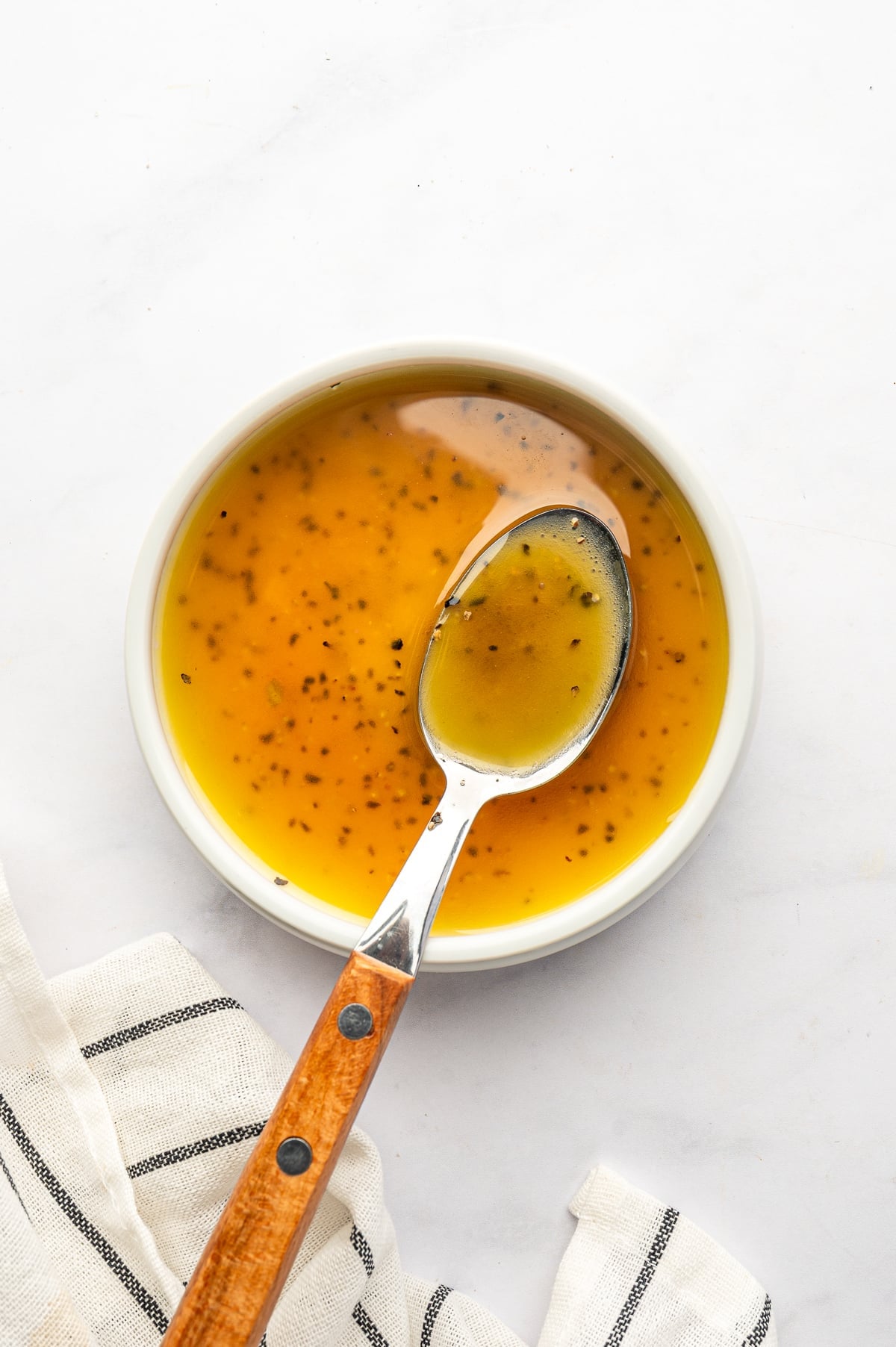 Golden olive oil and red wine vinegar dressing flecked with black pepper in a small white bowl, with a silver spoon with a wooden handle and a white towel with thin black stripes.