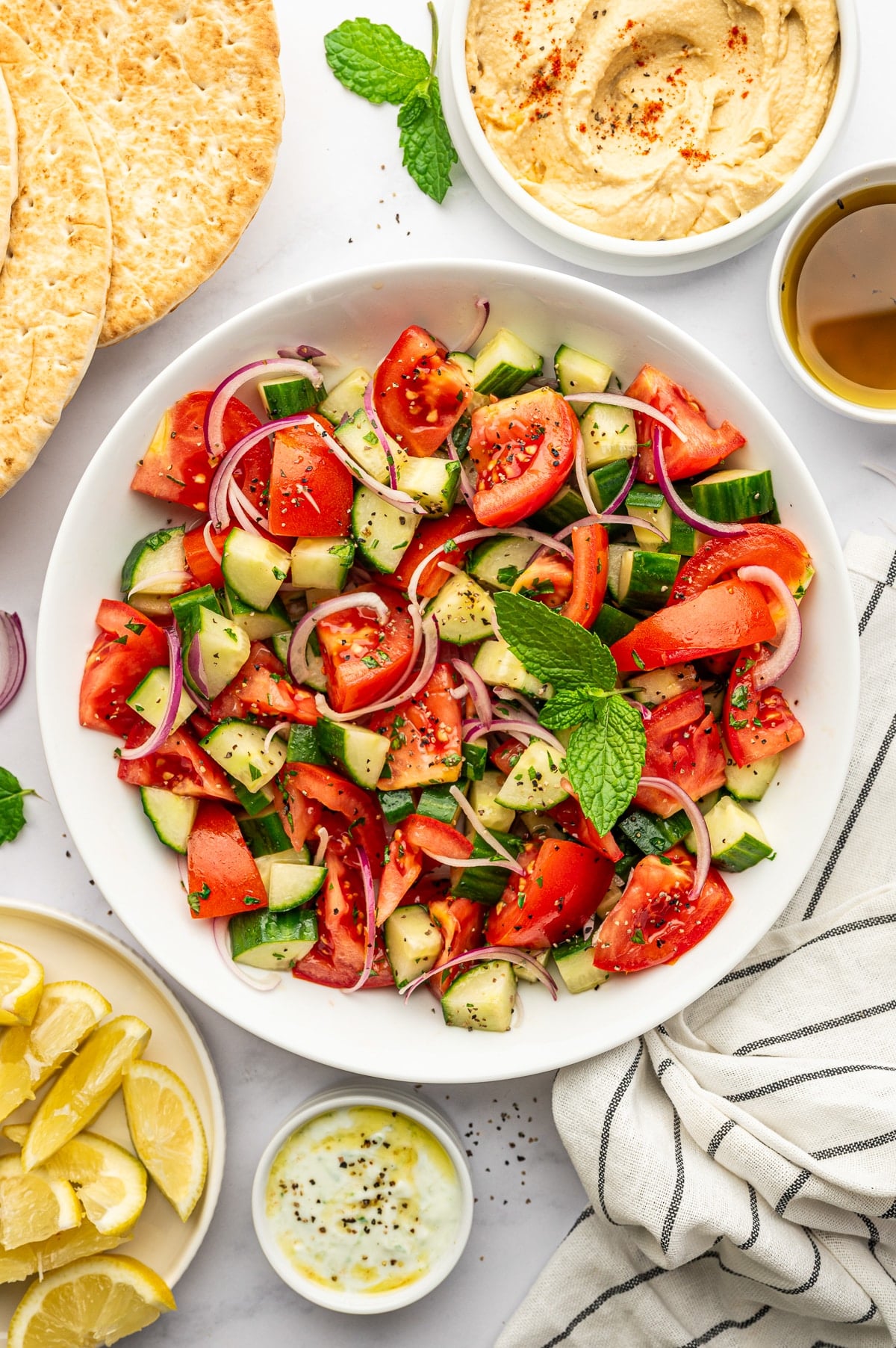 Cucumber tomato salad in a large white serving bowl with red tomato wedges, chopped green cucumber, thinly sliced red onion, parsley, black pepper, and a fresh mint garnish, surrounded by pita bread, hummus, lemon wedges, vinaigrette, and a white striped towel.
