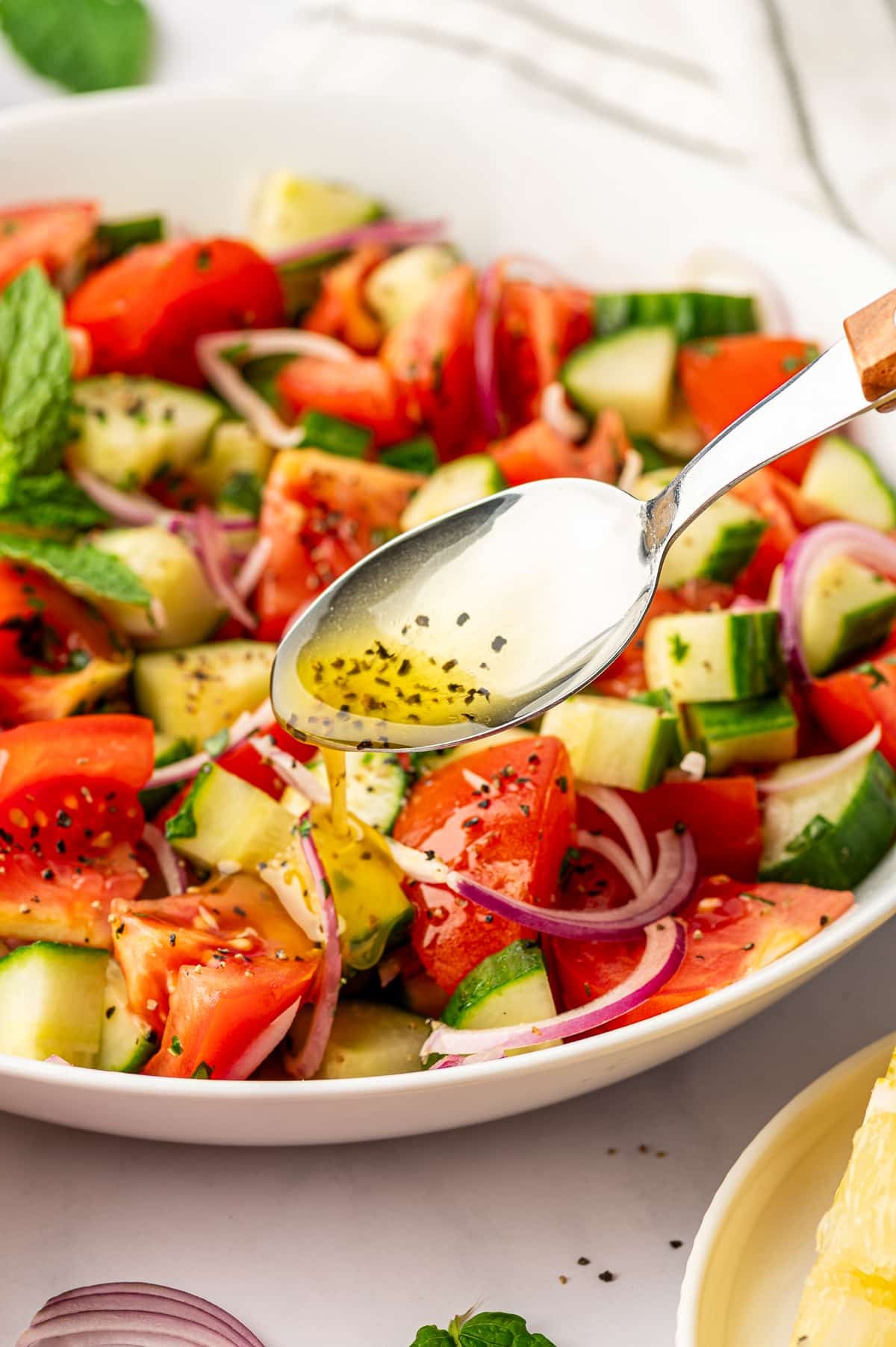 A silver spoon with a wooden handle drizzling golden vinaigrette over cucumber tomato salad in a large white bowl filled with red tomato wedges, chopped green cucumber, sliced red onion, parsley, black pepper, and fresh mint.