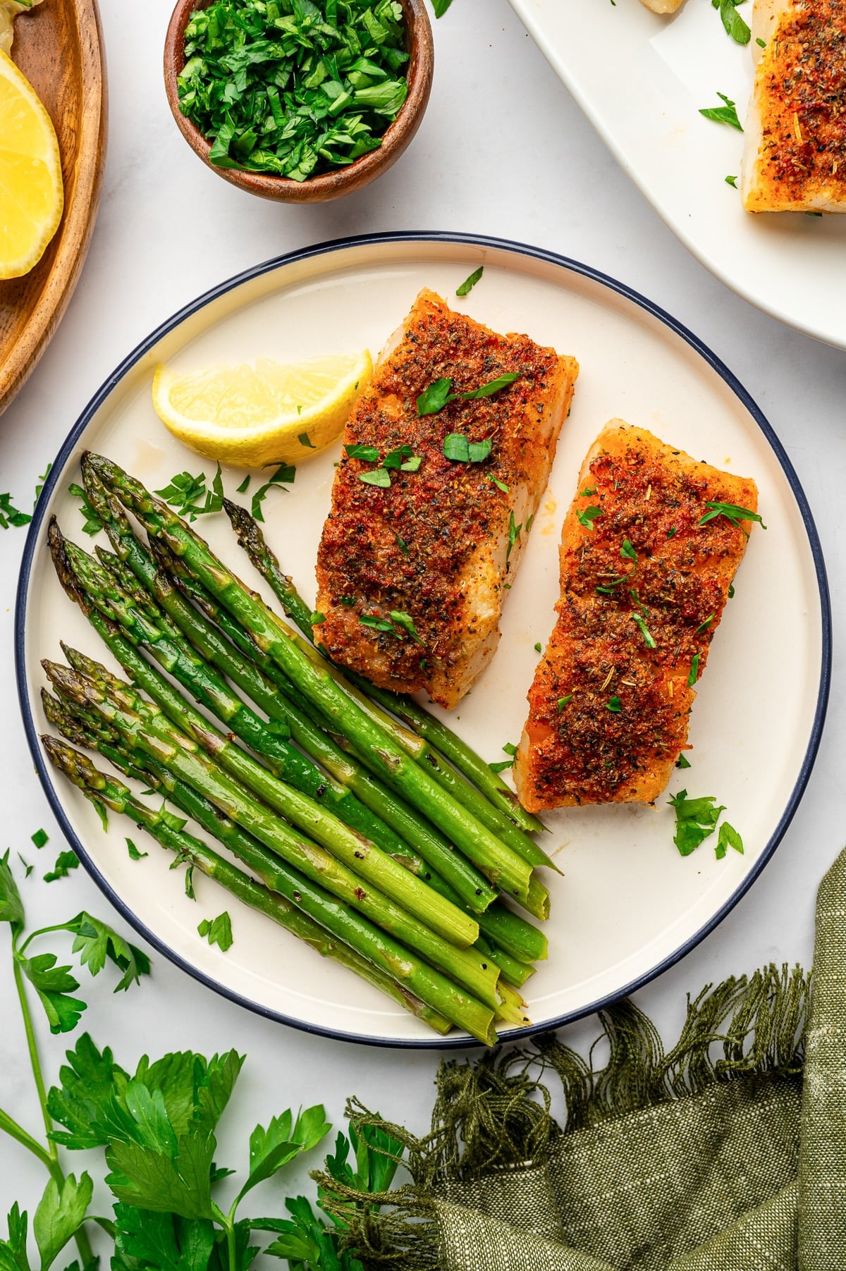 Overhead view of two paprika and herb-seasoned baked cod fillets with roasted asparagus on a cream-colored plate with a navy rim. The plate is garnished with parsley and a lemon wedge and surrounded by fresh parsley, chopped herbs, lemon, a white serving platter, and an olive-green fringed napkin.