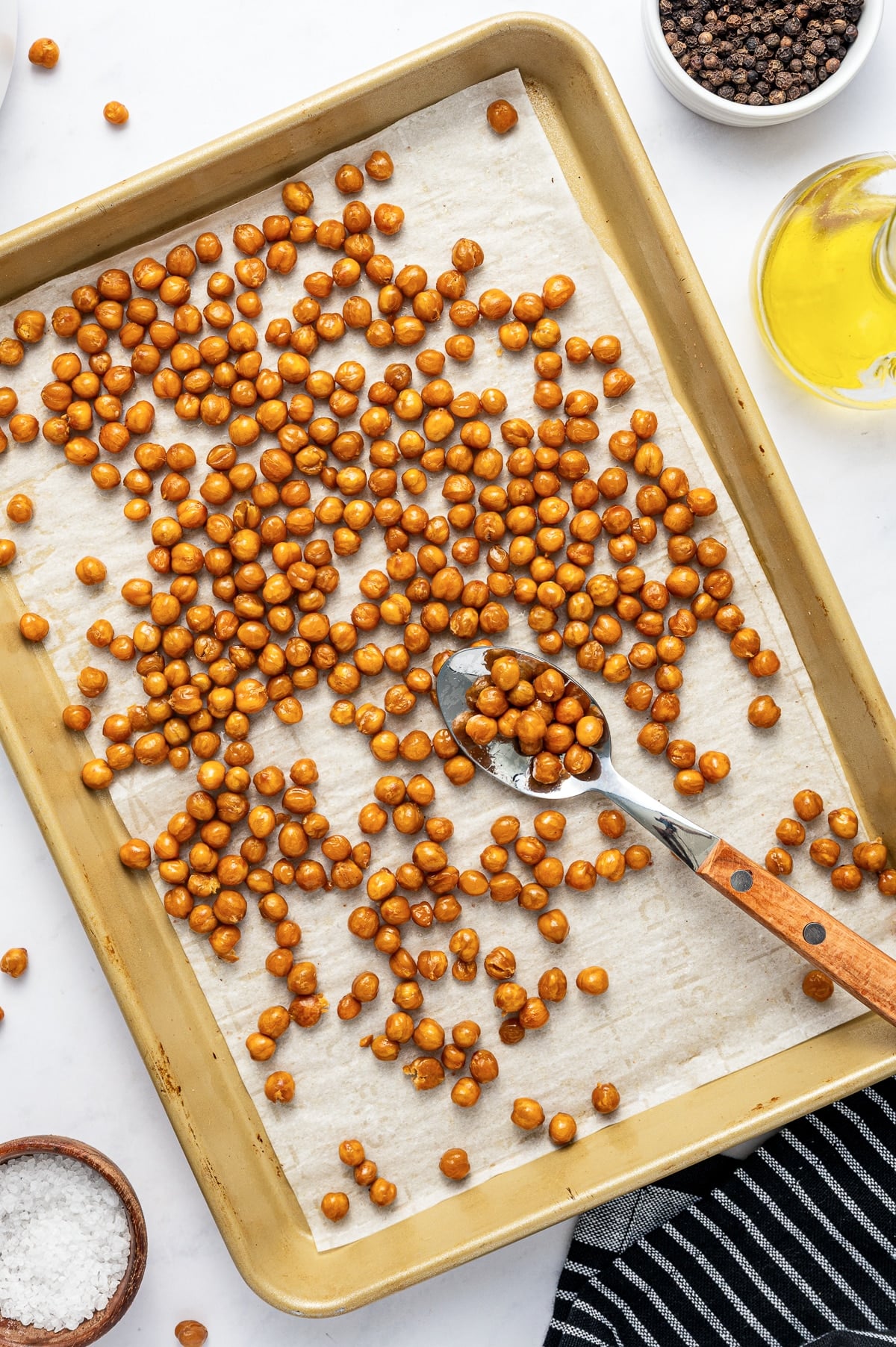 Golden oven-roasted chickpeas spread over white parchment paper on a gold-colored baking sheet with a wooden-handled silver spoon, a clear glass bottle of olive oil, a white bowl of black peppercorns, a wooden bowl of coarse salt, and a black-and-white striped towel.