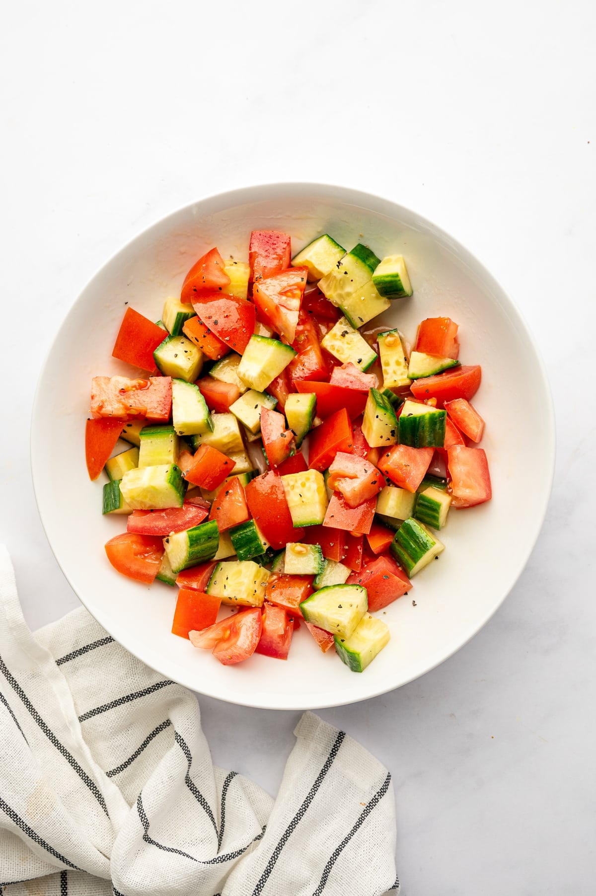 Fresh cucumber and red tomato chunks tossed with a light vinaigrette and black pepper in a shallow white bowl, photographed from overhead beside a loosely folded white towel with black stripes.