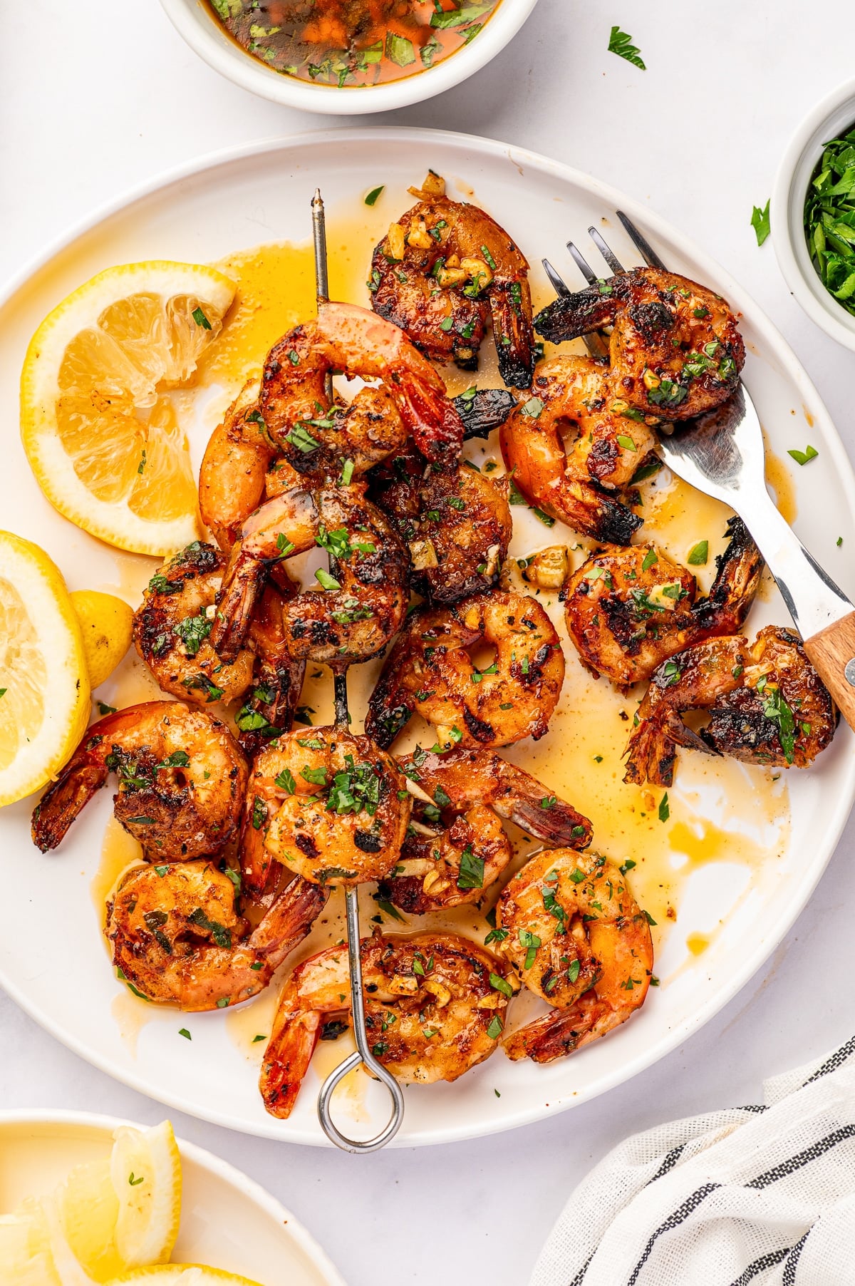 Overhead view of grilled shrimp kabobs on a large white plate with chopped parsley, minced garlic, golden marinade, and lemon wedges. A silver fork rests on the plate beside several partially served shrimp skewers.