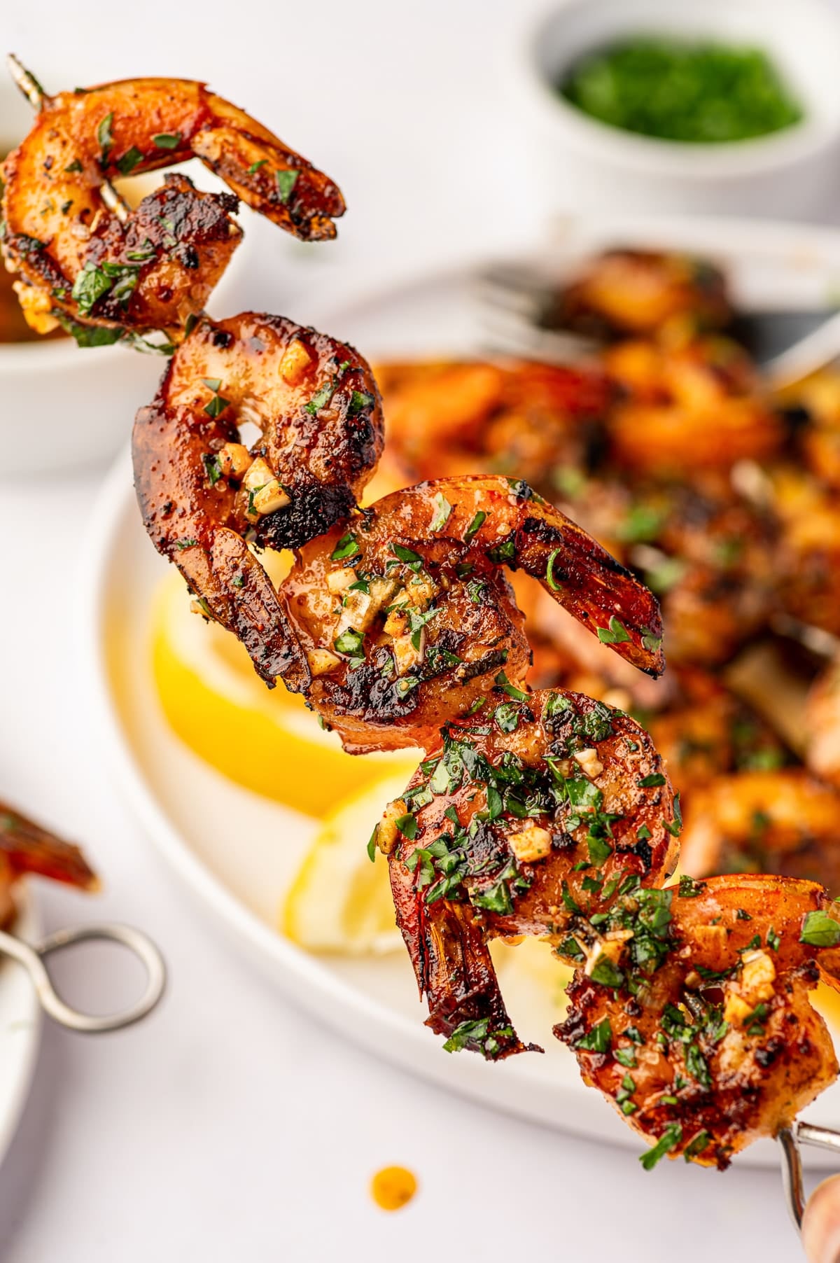 Close-up of a silver metal skewer holding several charred grilled shrimp coated with chopped garlic, fresh parsley, paprika, and glossy lemon marinade. A platter of additional shrimp and lemon wedges is softly blurred in the background.