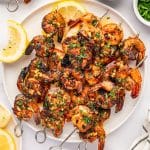 Overhead view of a large white platter filled with grilled shrimp skewers coated in lemon garlic marinade and garnished with chopped parsley. Lemon wedges, a bowl of marinade, a bowl of parsley, and a white towel with black stripes surround the platter.
