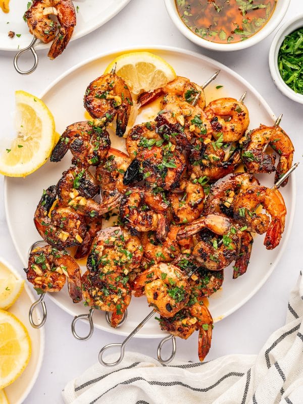 Overhead view of a large white platter filled with grilled shrimp skewers coated in lemon garlic marinade and garnished with chopped parsley. Lemon wedges, a bowl of marinade, a bowl of parsley, and a white towel with black stripes surround the platter.