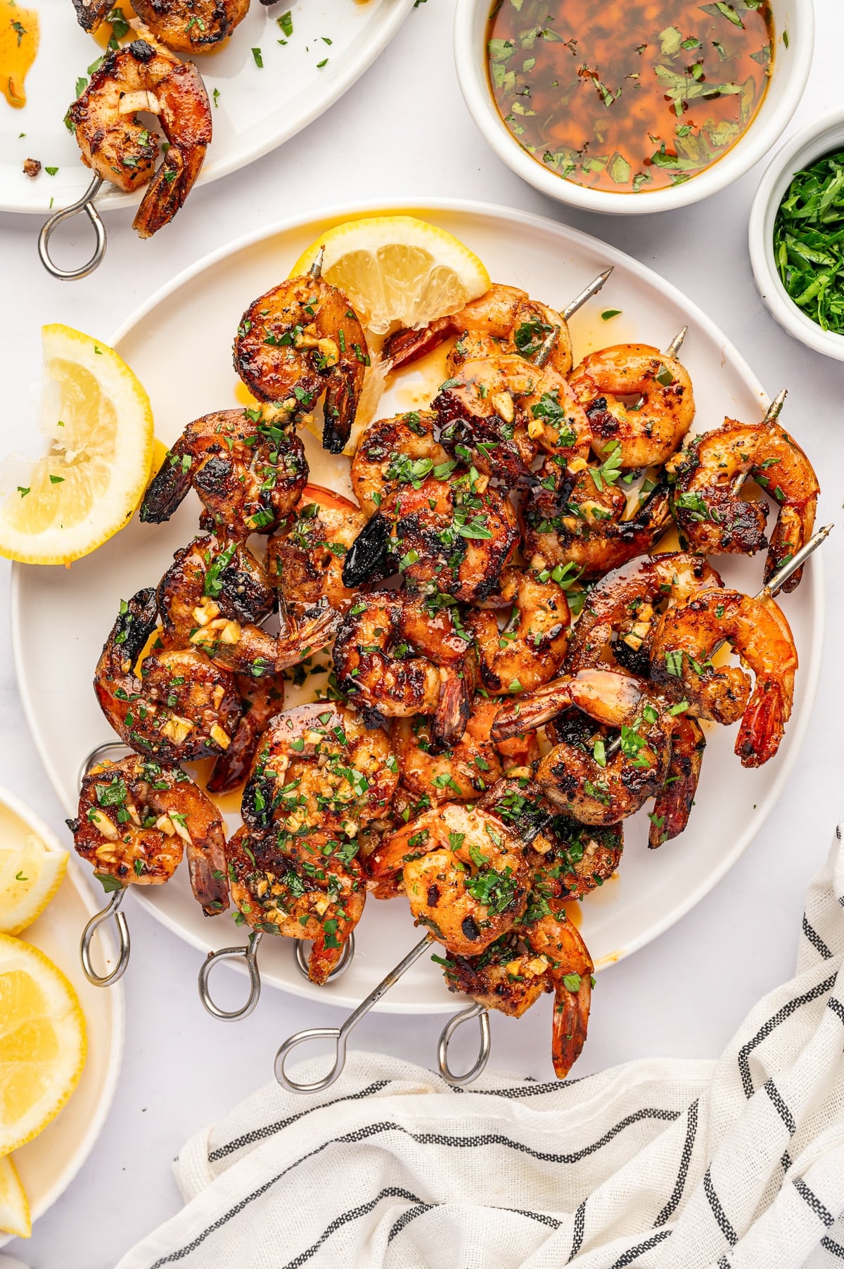 Overhead view of a large white platter filled with grilled shrimp skewers coated in lemon garlic marinade and garnished with chopped parsley. Lemon wedges, a bowl of marinade, a bowl of parsley, and a white towel with black stripes surround the platter.