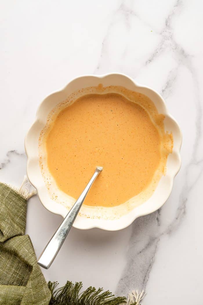 Overhead view of smooth, pale orange spicy mayo sauce in an ivory scalloped ceramic bowl with a silver spoon resting inside. Fine red seasoning flecks are visible throughout the creamy sauce, and an olive-green fringed cloth rests beside the bowl on a white marble background. No