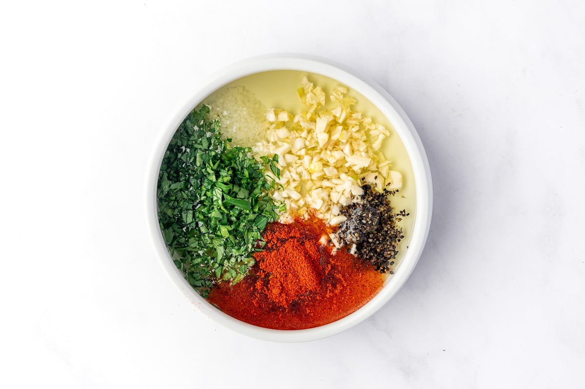 Overhead view of olive oil, finely chopped garlic, paprika, chopped fresh parsley, kosher salt, and cracked black pepper arranged separately in a small white bowl on a white marble surface.