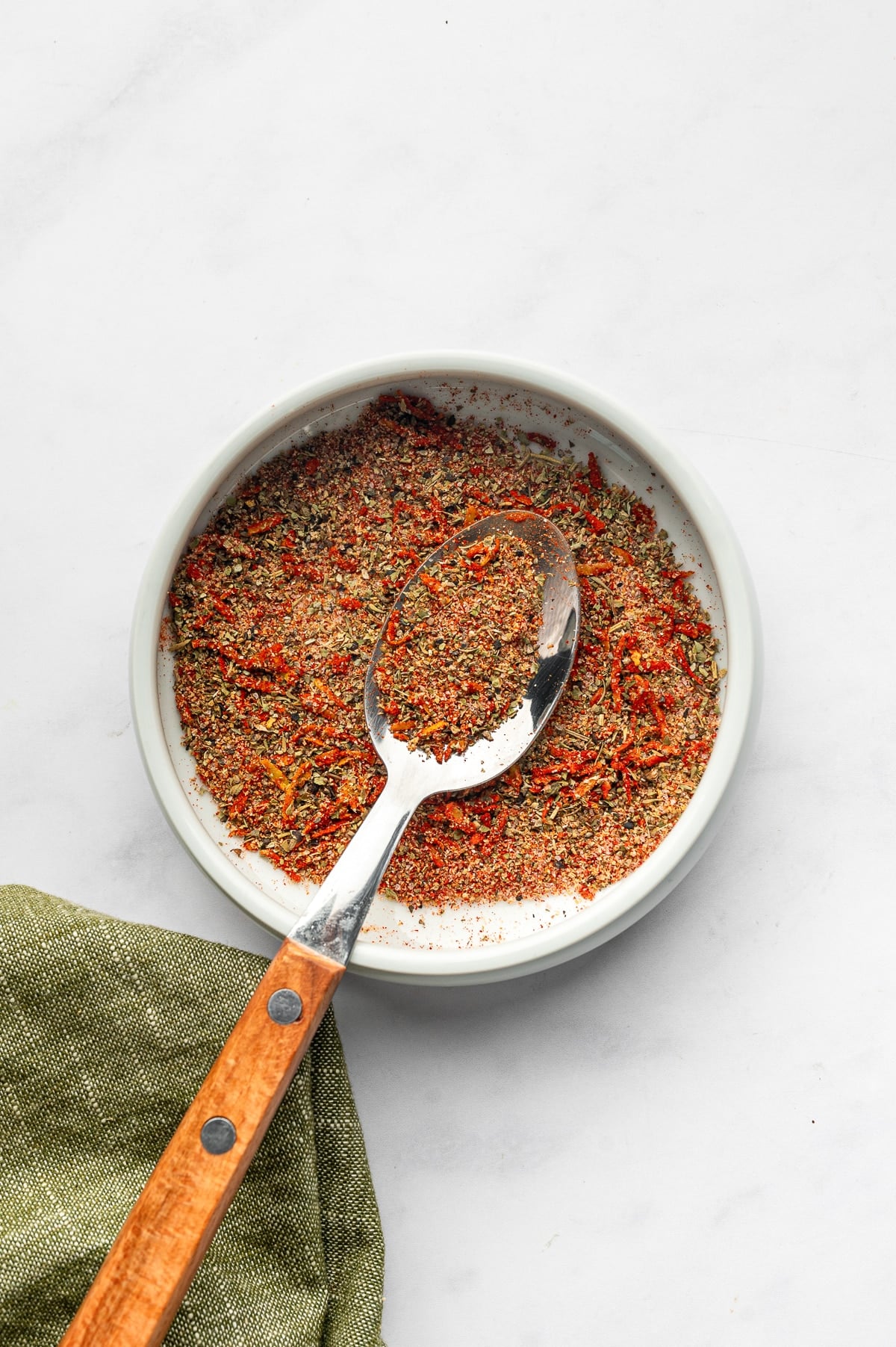 Mixed lemon-herb seasoning for baked cod in a shallow pale gray ceramic bowl with a silver spoon and warm brown wooden handle. An olive-green cloth napkin rests along the lower-left edge.