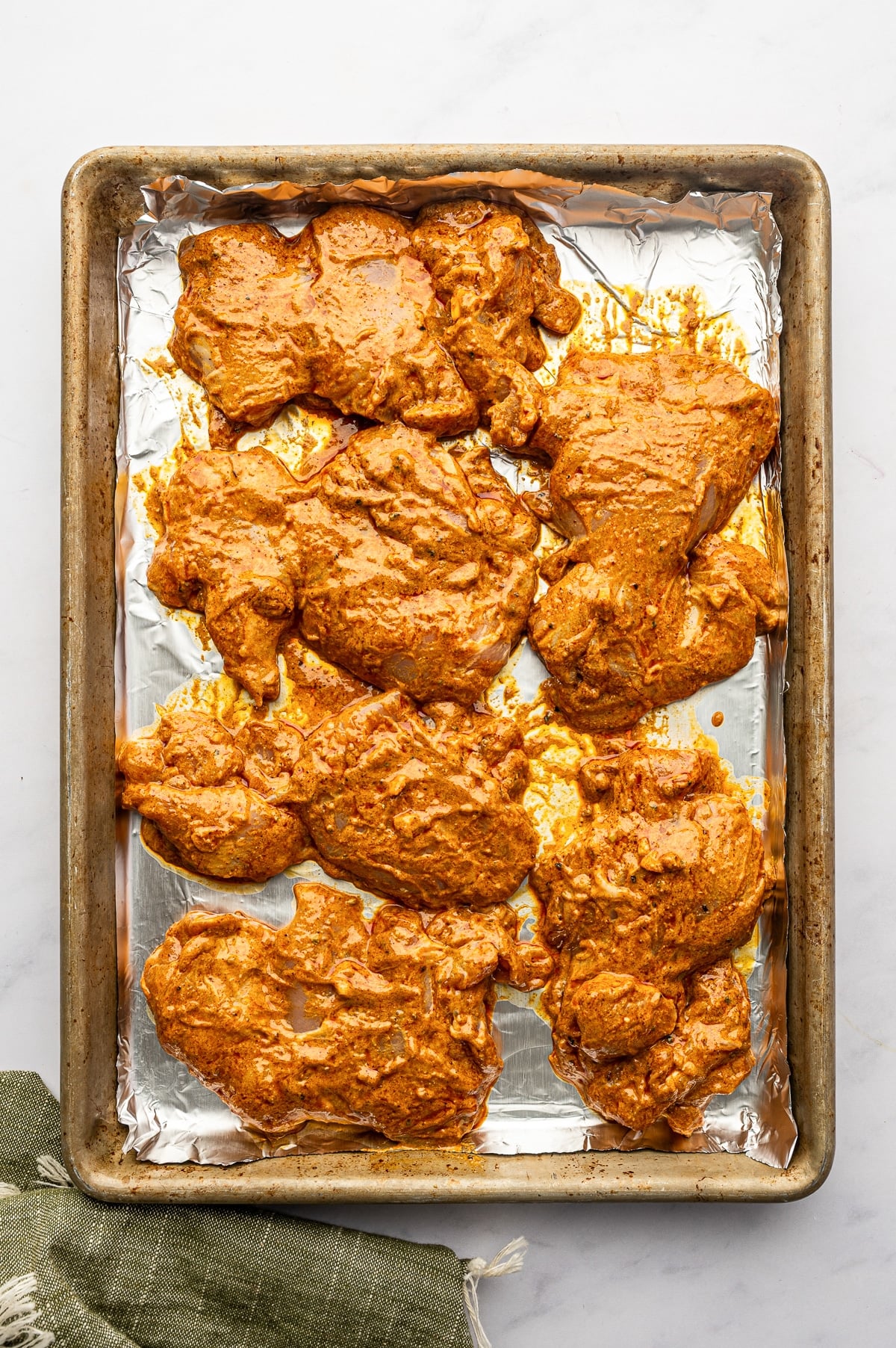 Raw marinated boneless chicken thighs arranged in a single layer on a foil-lined metal rimmed baking sheet, with a thick orange shawarma marinade coating each piece.