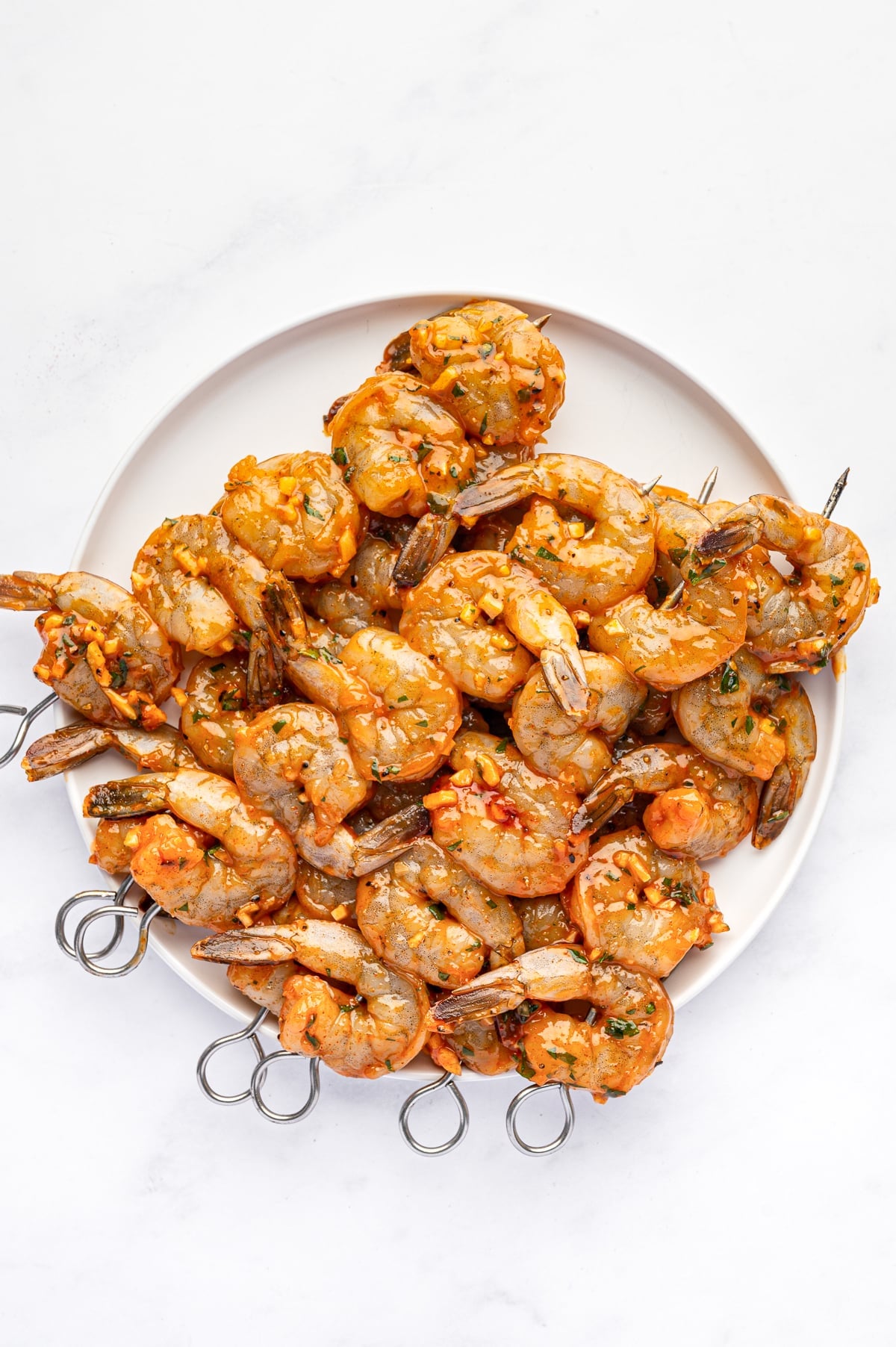 Overhead view of raw marinated shrimp threaded onto silver metal skewers and piled on a round white plate. The shrimp are coated with olive oil, garlic, paprika, parsley, and black pepper.