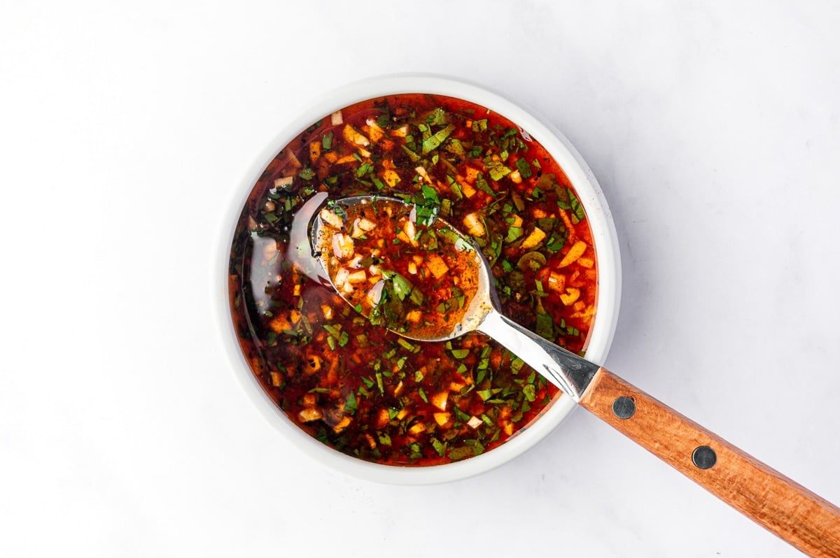 Overhead view of a red-orange lemon garlic shrimp marinade with chopped parsley, minced garlic, paprika, black pepper, and olive oil in a small white bowl with a silver spoon and wooden handle.