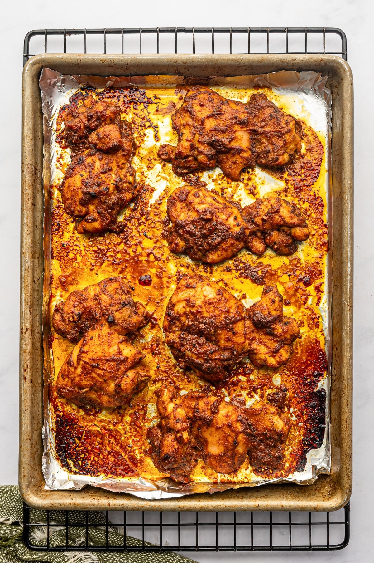 Deeply browned oven-baked chicken shawarma thighs on a foil-lined silver baking sheet set over a black cooling rack, surrounded by caramelized orange-red pan juices and charred seasoning.