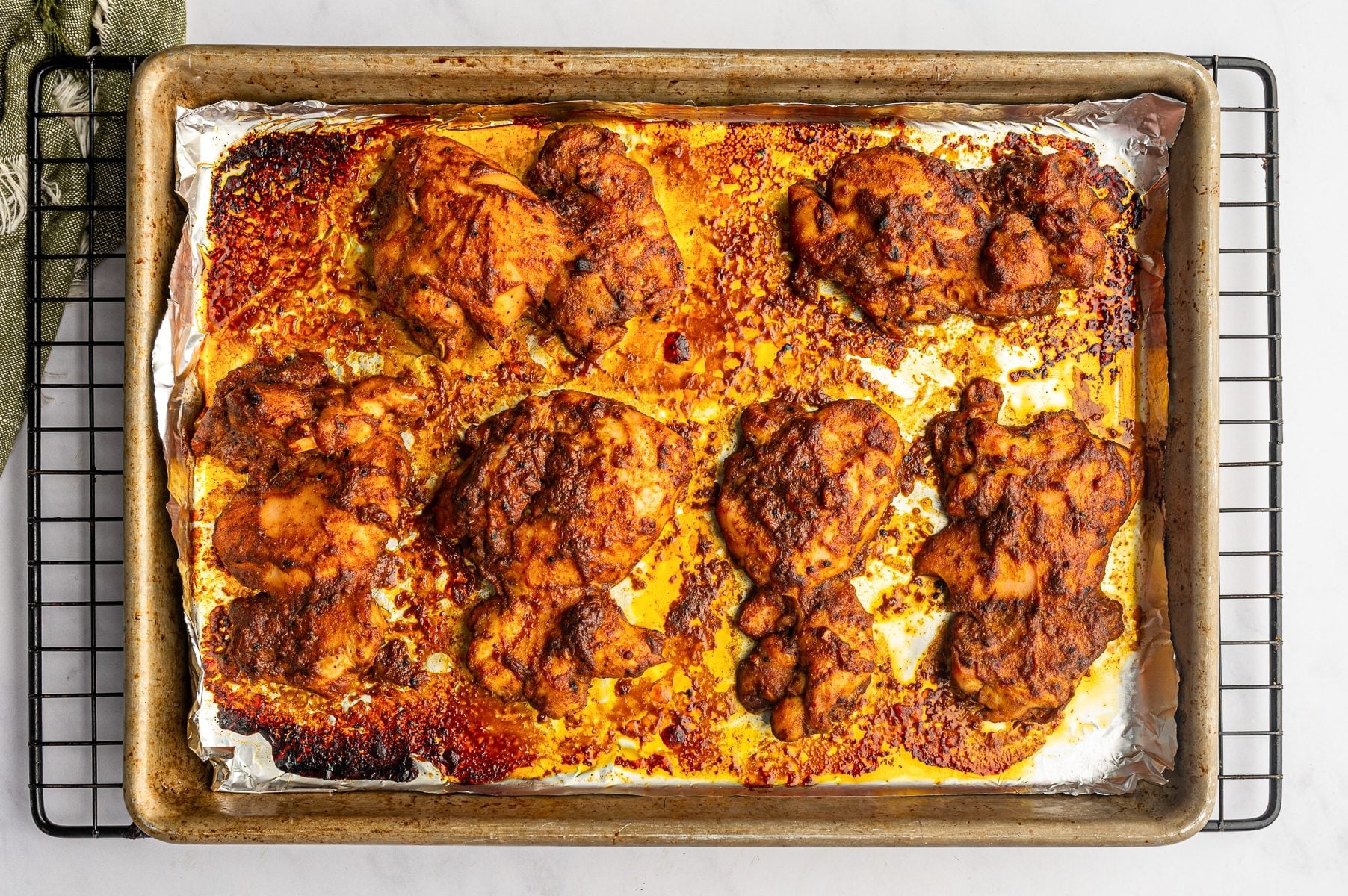 Oven-baked chicken shawarma thighs with deeply browned and lightly charred edges on a foil-lined metal sheet pan set over a black cooling rack, surrounded by orange pan juices and caramelized marinade.
