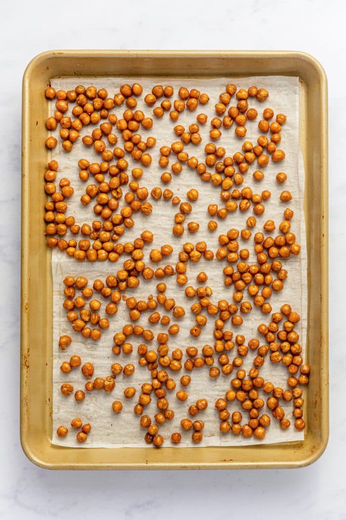 Golden-brown oven-roasted chickpeas spread across white parchment paper inside a gold-colored rimmed baking sheet, photographed overhead after roasting.