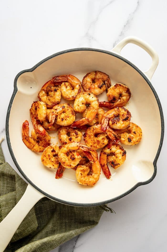 Overhead view of seasoned shrimp with tails on, cooked until golden-orange with browned and lightly charred edges in a large cream-colored enameled skillet with a dark gray rim and cream handles. The skillet rests on a white marble surface over an olive-green fringed cloth.