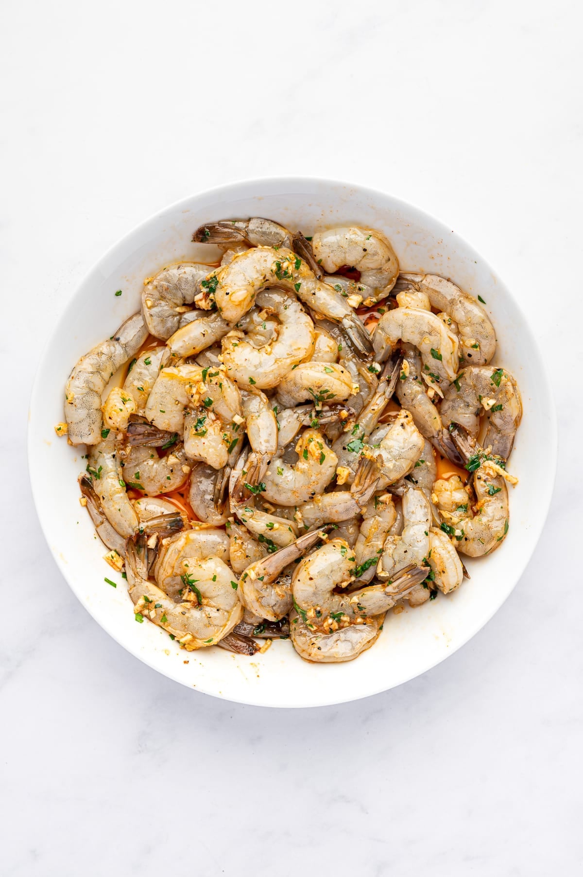 Overhead view of raw peeled and deveined shrimp with the tails attached, coated in a lemon garlic marinade with chopped parsley and seasonings in a large white bowl on a white marble surface.