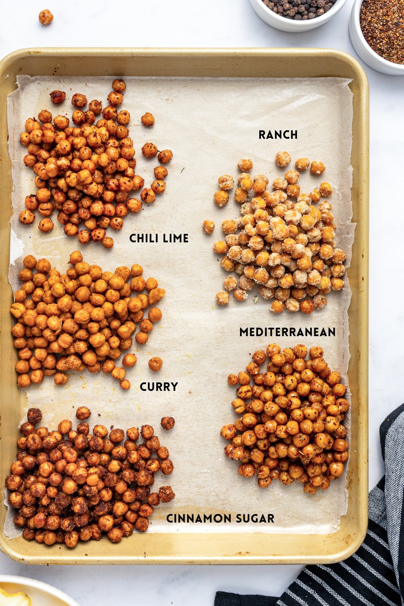 Five piles of roasted chickpeas arranged on a parchment-lined gold baking sheet with black text labels identifying each flavor: Chili Lime at the upper left, Ranch at the upper right, Curry at the middle left, Mediterranean at the lower right, and Cinnamon Sugar at the lower left.