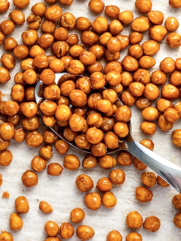 Golden roasted chickpeas scattered over white parchment paper with a large silver spoon filled with chickpeas positioned diagonally across the center.