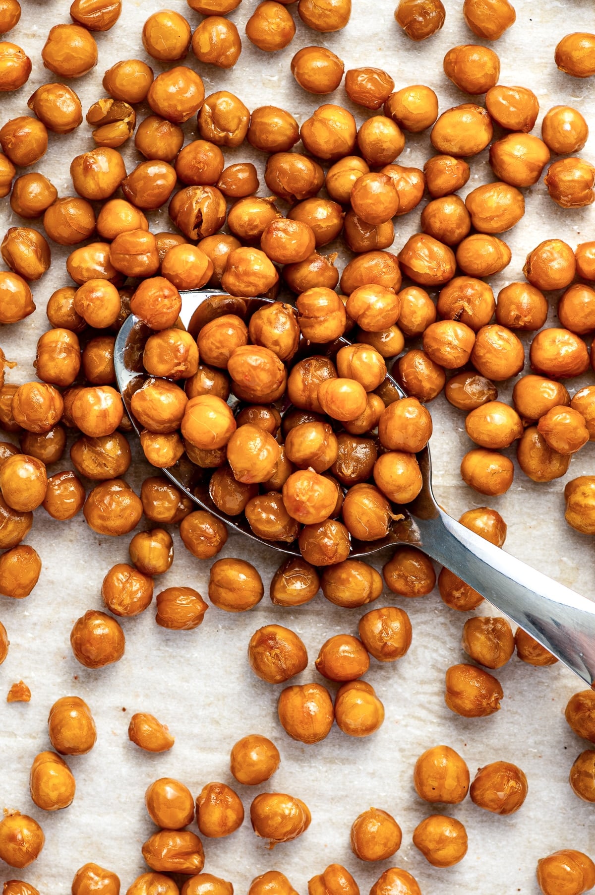 Golden roasted chickpeas scattered over white parchment paper with a large silver spoon filled with chickpeas positioned diagonally across the center.