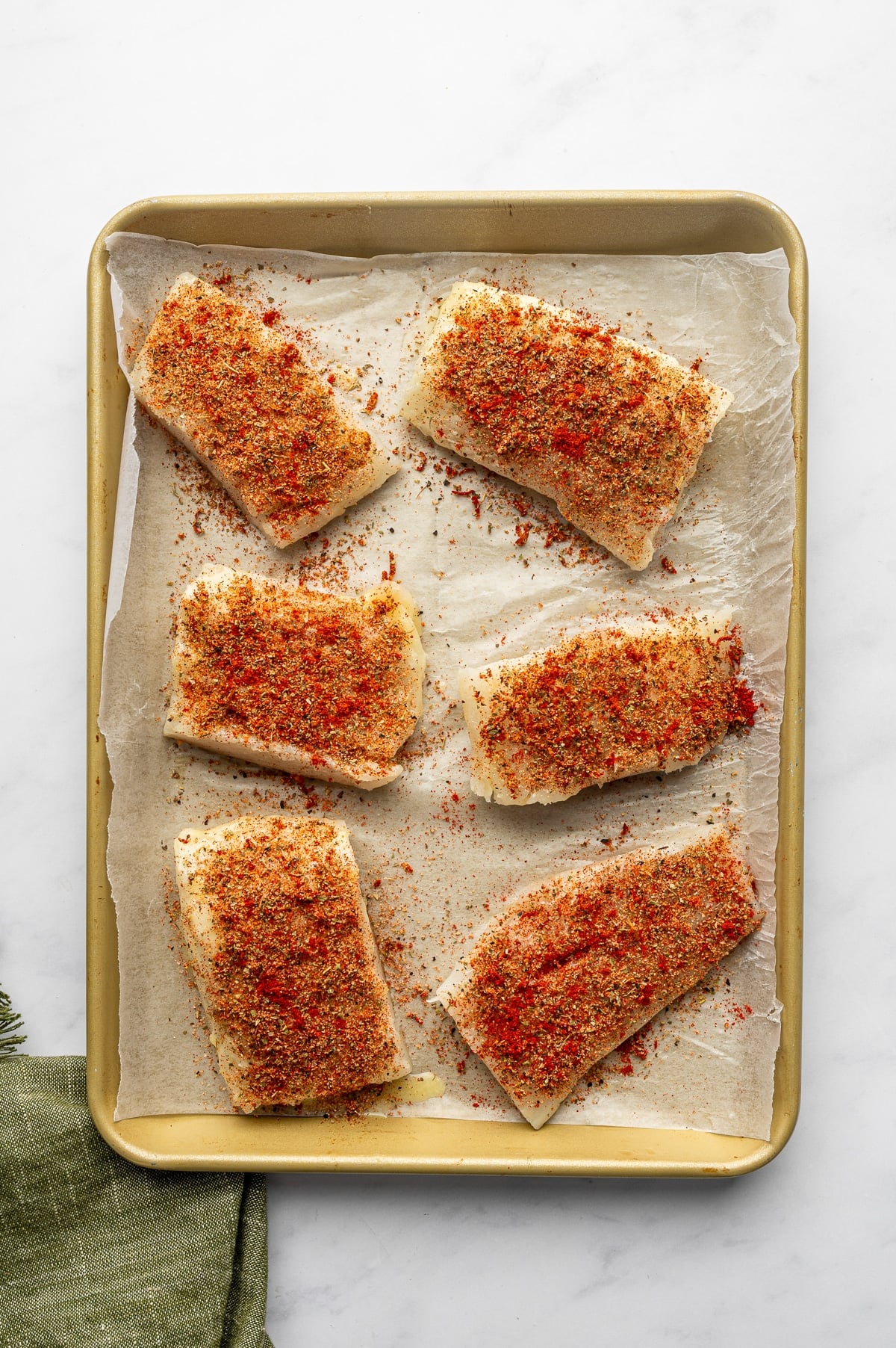 Six raw cod fillets coated with a red and green lemon-herb seasoning blend on tan parchment paper inside a gold rimmed baking sheet. An olive-green cloth napkin appears at the lower-left edge.