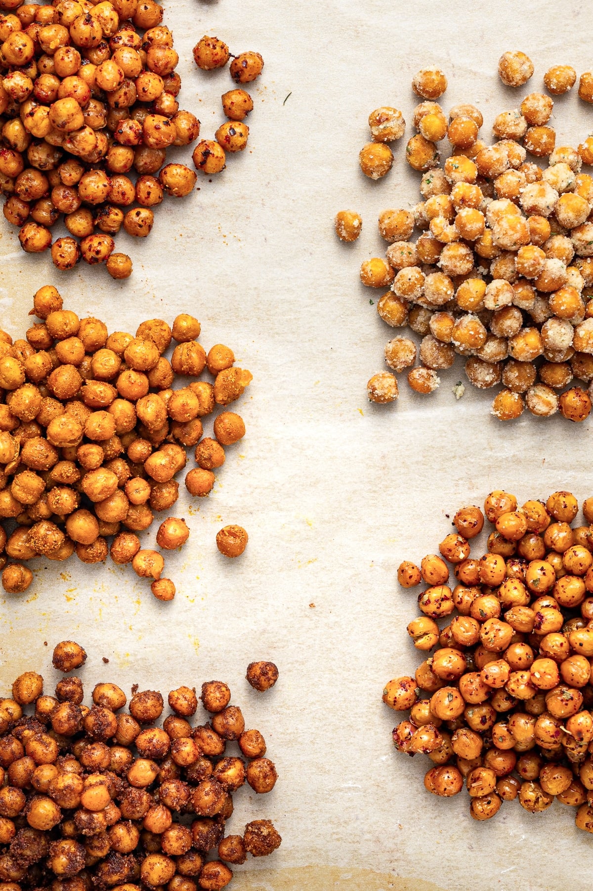 Close overhead view of five roasted chickpea variations arranged around the edges of white parchment paper, including red-speckled chili lime, pale ranch-coated, golden curry, herb-speckled Mediterranean, and dark cinnamon sugar chickpeas.