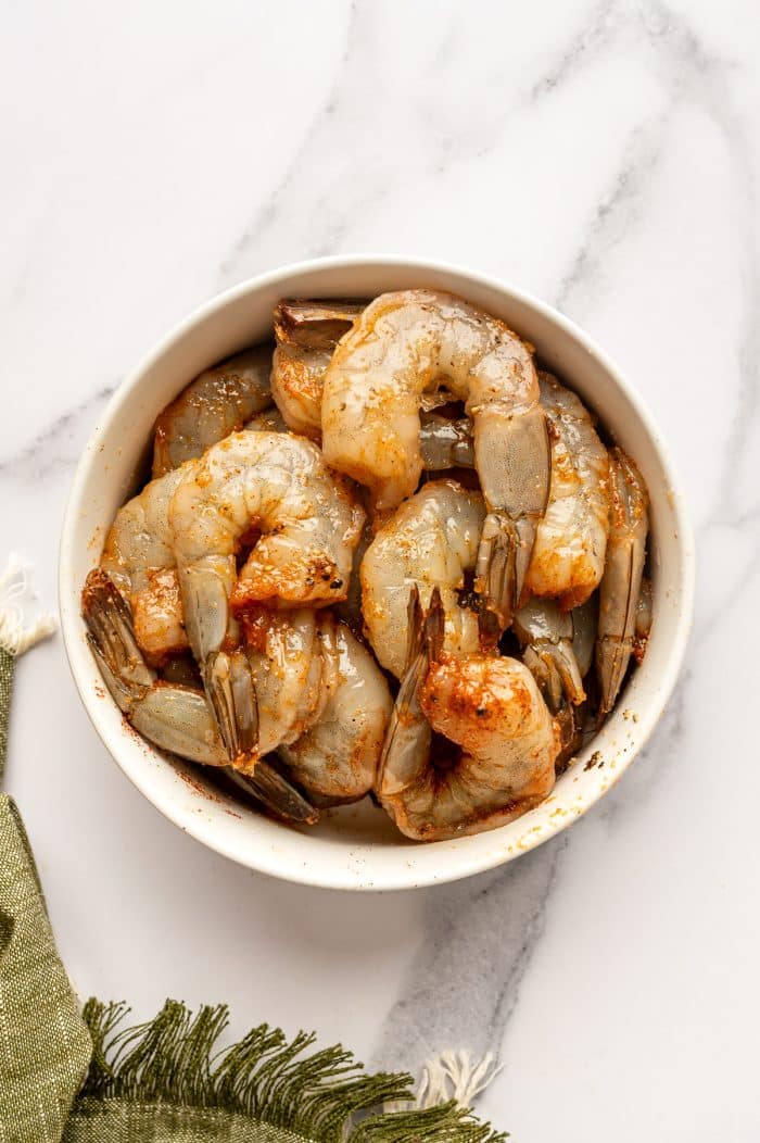 Overhead view of raw shrimp with tails on tossed with oil, paprika, garlic powder, onion powder, salt, and black pepper in a round white ceramic bowl. The coated shrimp have a light orange-red color and sit on a white marble surface beside an olive-green fringed cloth.