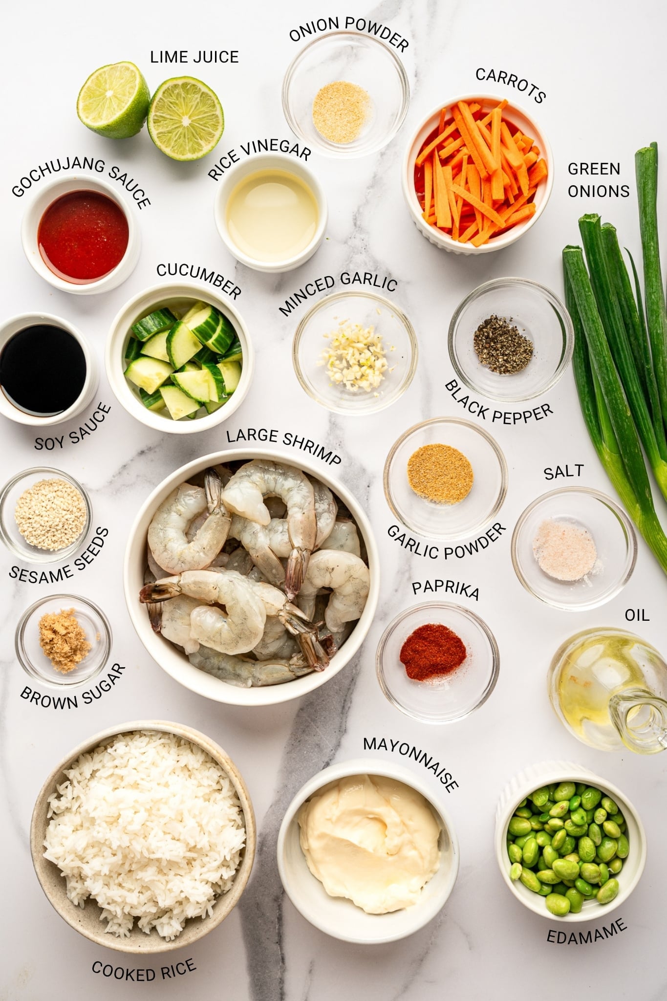Overhead ingredient layout for a shrimp rice bowl arranged on a white marble surface. Ingredients include raw shrimp in a white bowl, cooked white rice, mayonnaise, edamame, matchstick carrots, diced cucumber, green onions, lime juice, gochujang sauce, soy sauce, rice vinegar, minced garlic, brown sugar, oil, paprika, garlic powder, onion powder, salt, black pepper, and sesame seeds. Black text overlays identify each ingredient with the labels "LIME JUICE," "ONION POWDER," "CARROTS," "GOCHUJANG SAUCE," "RICE VINEGAR," "GREEN ONIONS," "CUCUMBER," "MINCED GARLIC," "BLACK PEPPER," "SOY SAUCE," "LARGE SHRIMP," "GARLIC POWDER," "SALT," "SESAME SEEDS," "BROWN SUGAR," "PAPRIKA," "OIL," "COOKED RICE," "MAYONNAISE," and "EDAMAME."