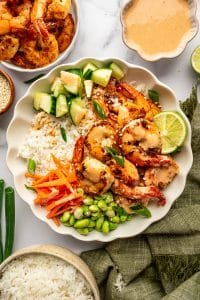 Overhead view of a complete shrimp rice bowl in a large ivory scalloped bowl filled with white jasmine rice, golden pan-seared shrimp, diced cucumber, matchstick carrots, and bright green edamame. The shrimp are drizzled with creamy orange spicy mayo and garnished with sliced green onions, white sesame seeds, and a lime wedge. A white bowl of extra shrimp, an ivory bowl of spicy mayo, a small brown bowl of sesame seeds, and a beige bowl of rice surround the main bowl on a white marble surface with an olive-green fringed cloth.