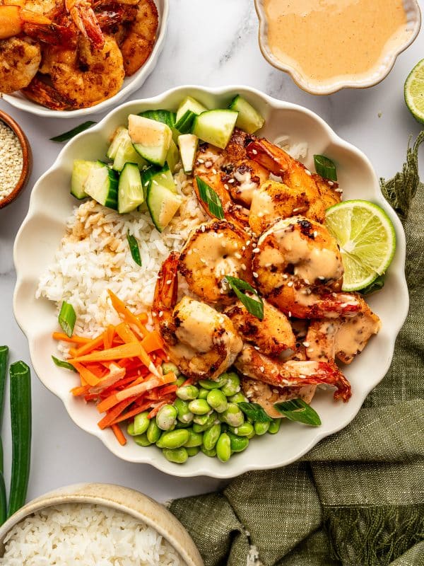 Overhead view of a complete shrimp rice bowl in a large ivory scalloped bowl filled with white jasmine rice, golden pan-seared shrimp, diced cucumber, matchstick carrots, and bright green edamame. The shrimp are drizzled with creamy orange spicy mayo and garnished with sliced green onions, white sesame seeds, and a lime wedge. A white bowl of extra shrimp, an ivory bowl of spicy mayo, a small brown bowl of sesame seeds, and a beige bowl of rice surround the main bowl on a white marble surface with an olive-green fringed cloth.
